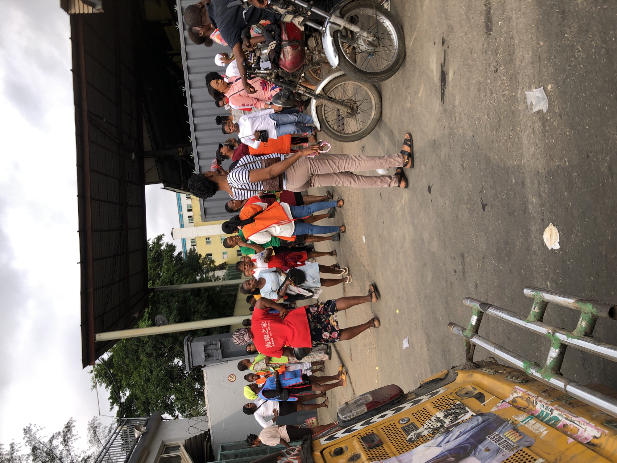 The image depicts a street scene in Lagos, Nigeria, with a group of people gathered outdoors. A yellow motorized tricycle, commonly known as a Keke Napep, is partially visible on the left side of the frame, adorned with stickers. In the background, a low-lying building with a corrugated iron roof overhang extends across the scene, supported by metal poles. Several individuals, predominantly women, are standing and interacting. One woman in a red shirt with Chinese characters and a floral skirt is standing in the center, with her back to the camera. Other women are seen in various casual attire, some wearing vests or head coverings. Two motorcycles are parked near the center right of the image. The ground is asphalt, with some debris scattered on it. The sky above is overcast, suggesting a cloudy day. The overall atmosphere appears to be that of a bustling urban environment.