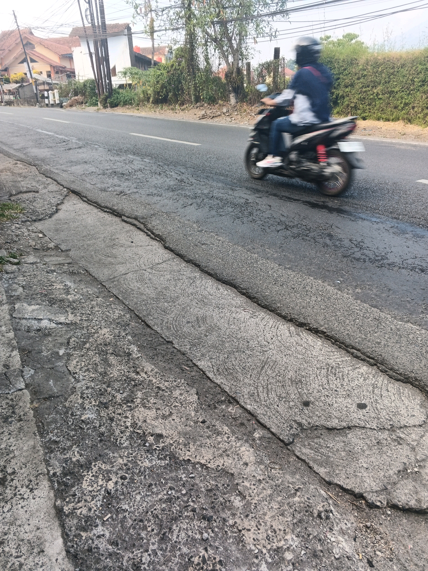 A motorcycle is speeding down a road in Ungaran, Indonesia. The rider is wearing a helmet and a dark blue jacket, and is blurry due to the speed. The road is asphalt, with some cracks and patches. There are buildings and trees on the side of the road. The sky is overcast.