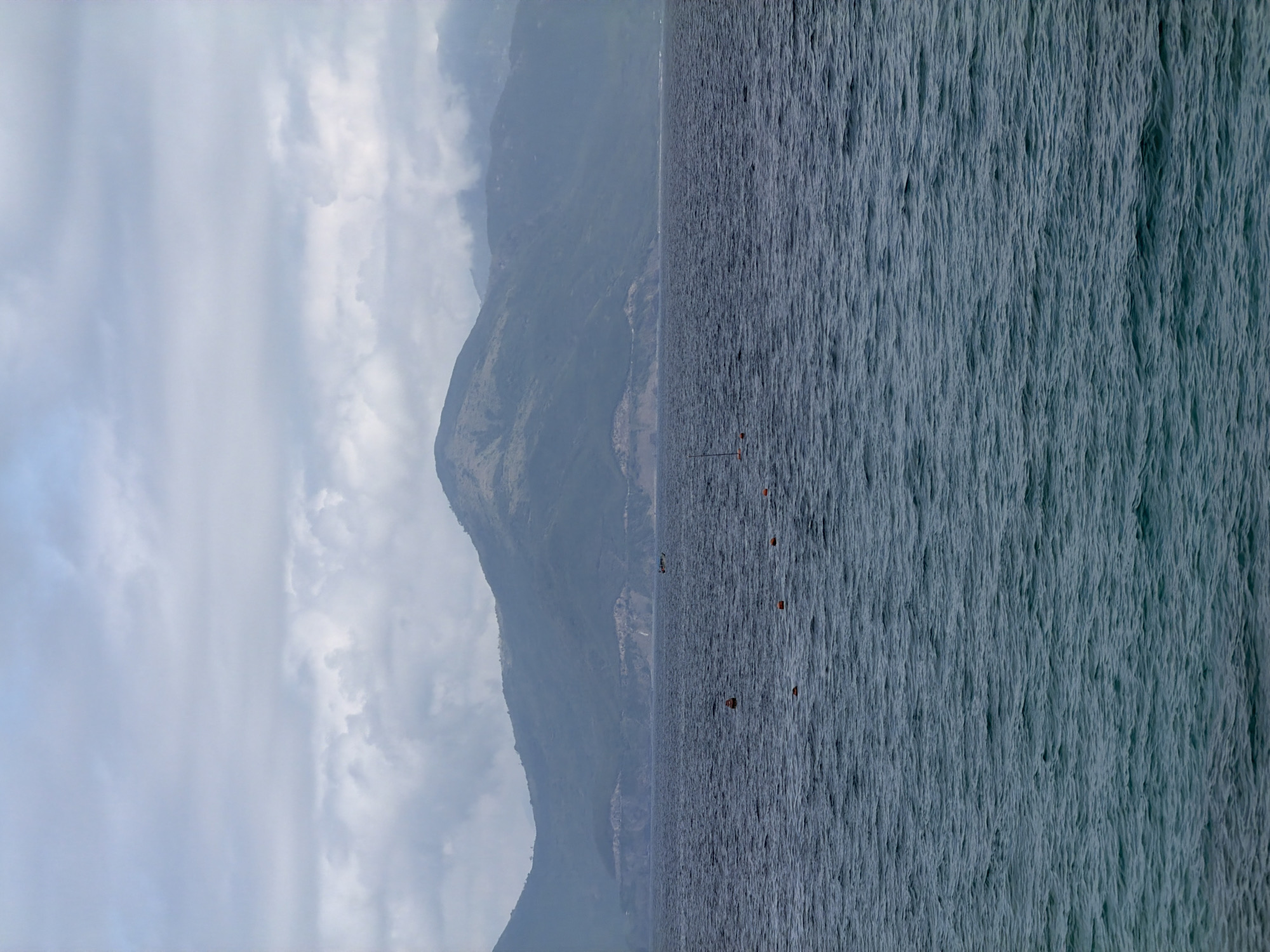 The image depicts a waterscape scene. On the right side, there is an expanse of water, textured with light waves. Several orange buoys are sparsely scattered across the water's surface. The left side of the image reveals a mountain range shrouded in mist and clouds. The scene appears to be captured from a high vantage point, looking out towards the sea and the distant mountains. While it shares characteristics with Nha Trang Ward, Vietnam, the image provides no distinct markers to confirm its precise location.