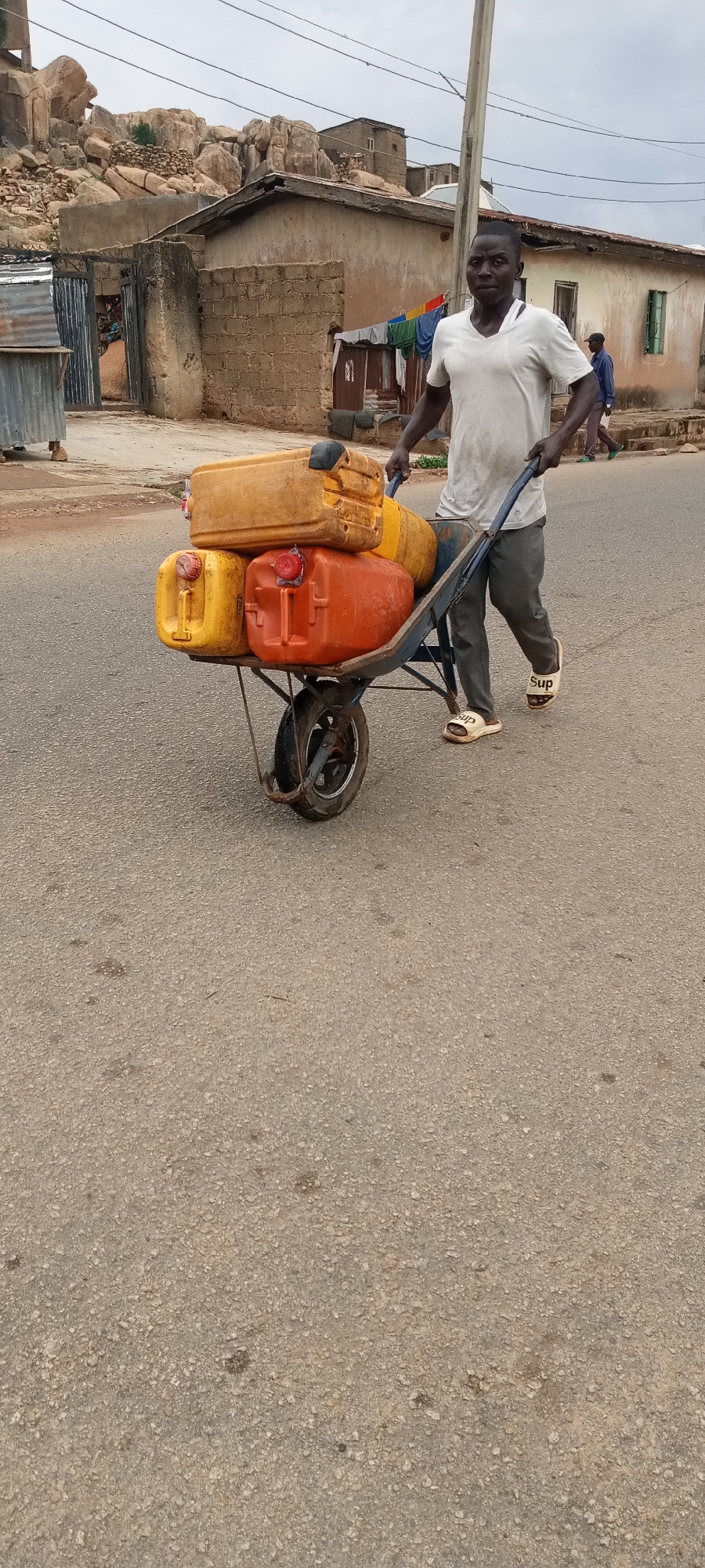 A man walks down a paved road in the city of Jos, Nigeria. He pushes a blue wheelbarrow loaded with four rectangular plastic containers, three yellow and one orange. He wears a white V-neck shirt, dark pants, and white sandals with "Sup" printed on them. In the background are buildings constructed of concrete and brick, along with a rock formation. Clothes hang on a line outside one building. Another man is walking in the background. The sky is overcast.