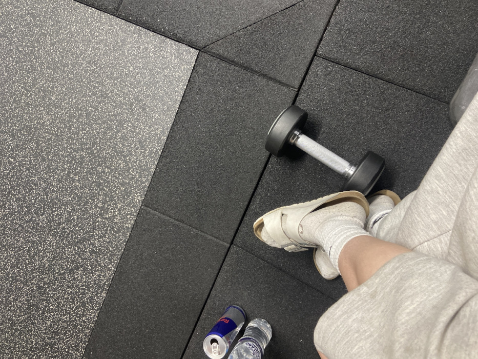 The image displays an overhead view of a person's lower body within what appears to be a gym or fitness center. The flooring consists of black interlocking rubber mats, with a diagonal section of speckled grey rubber matting visible in the upper right. The person is wearing light grey sweatpants, white ribbed ankle socks featuring a faint black Nike swoosh, and off-white or beige Birkenstock-style slide sandals. Their legs are crossed. On the black rubber mat, an open, partially consumed Red Bull energy drink can is present alongside a clear plastic bottle of still water, which has a visible "Highland" label, likely indicating "Highland Spring." A single black dumbbell with a silver knurled handle rests on the floor near the person's feet. The specific location within the city of London, United Kingdom, cannot be identified from the visual content provided.