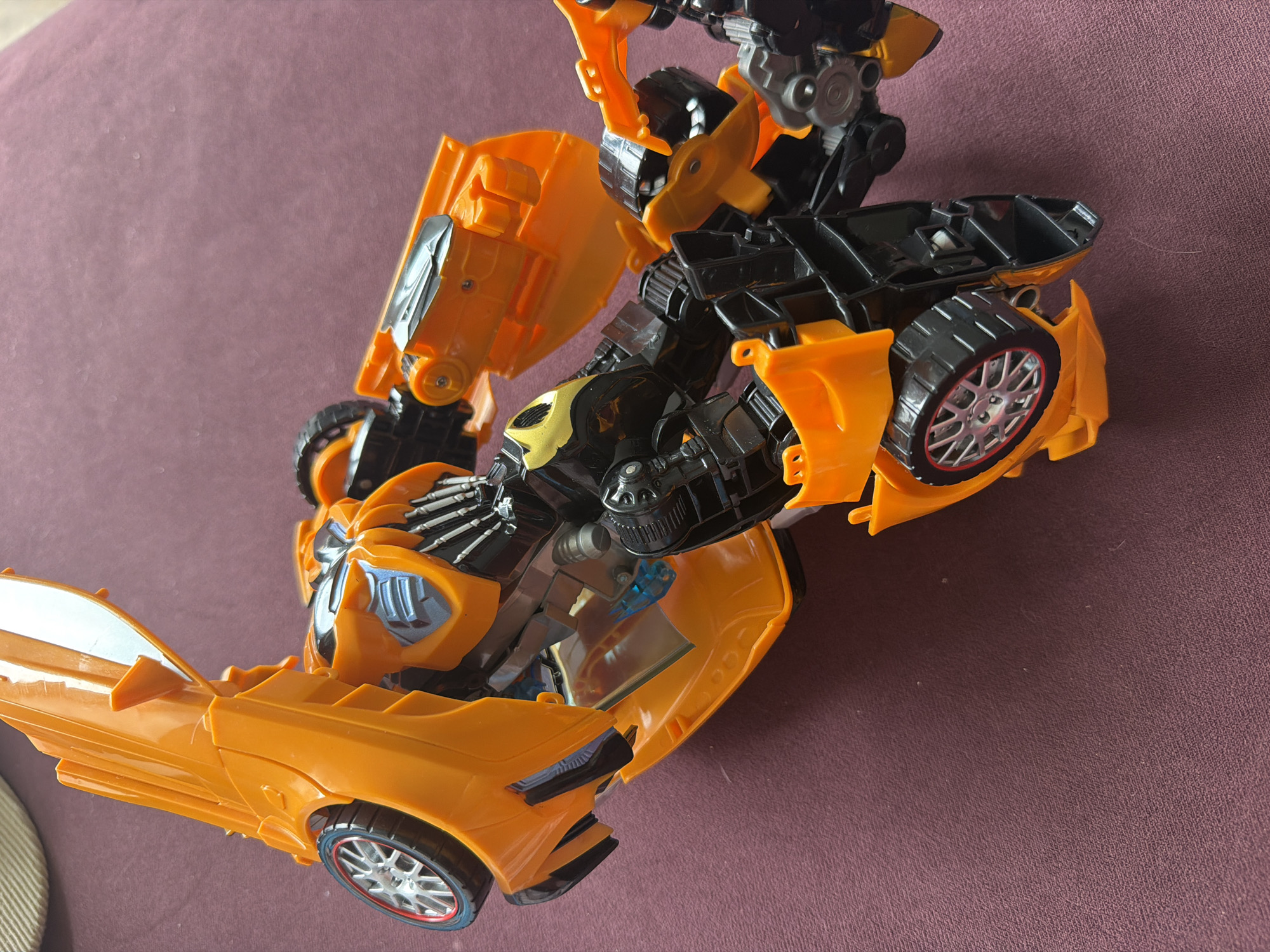 An orange and black transforming toy, resembling Bumblebee from Transformers, is depicted in a partially articulated state, midway between its vehicle and robot forms. The toy's primary color is a vibrant orange, complemented by significant black mechanical components and silver accents on its multi-spoke wheels and exposed internal structures. A small, translucent blue element is visible within the central chest area of the robot form. The toy lies diagonally on its right side, with sections of its car shell, including the roof/hood and rear panels, opened and angled upwards, revealing the internal robot limbs and torso. One black robot arm is extended towards the top of the frame, and the legs are bent underneath. The toy rests on a uniformly textured, dark reddish-brown fabric surface, likely carpet or a blanket. A light beige, out-of-focus object is partially visible in the upper left corner of the image. The image contains no discernible visual identifiers to establish a specific geographic location, including the city of Chaikovsky, Russia.