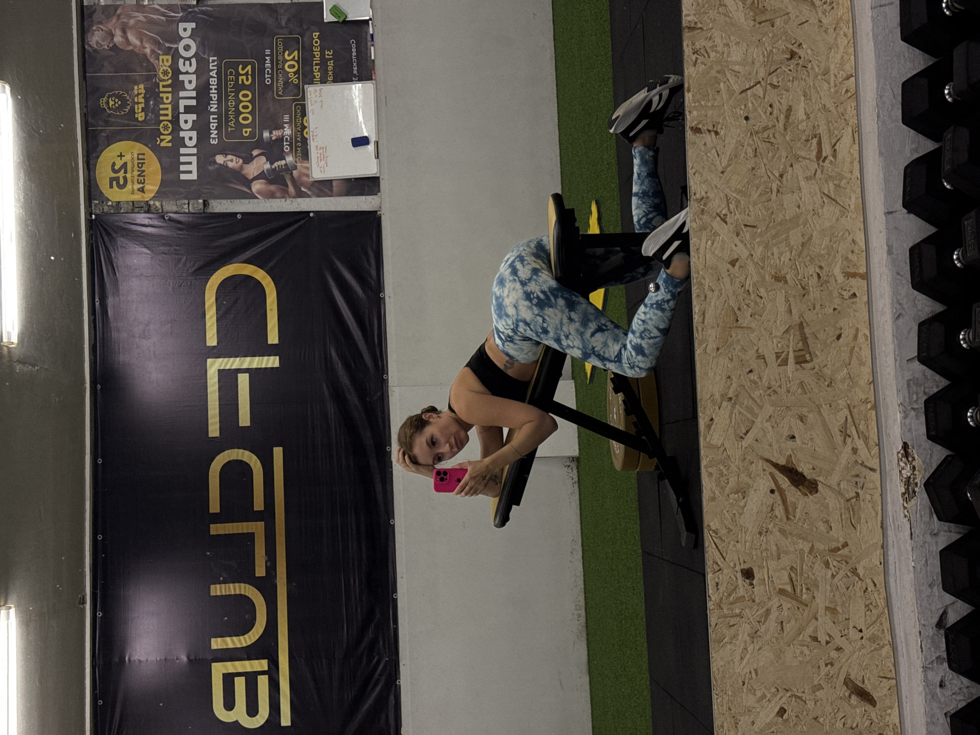 This media file captures a young woman posing in a fitness center in Chaikovsky, Russia. The image is taken indoors under artificial lighting. The woman, positioned on an exercise bench often used for hyperextensions, is facing the camera, holding a bright pink smartphone in her left hand. She wears a black sports bra and blue and white tie-dye patterned leggings with athletic shoes. Her expression is relaxed, with a slight smile.
The background features a concrete wall, with a large black banner displaying "CLUB" vertically in gold lettering. Above this, another banner advertises gym promotions, featuring images of a male and female bodybuilder. Visible text on this banner includes "50% СКИДКИ" (50% Discounts), "II МЕСЯЦ" (II Month), "СВОБОДНЫЙ ТРЕНЕР" (Free Trainer), "21 ДЕНЬ" (21 Days), "32 000 ₽" (32,000 Rubles), "ВЫБЕРИ БОЛЬШЕ" (Choose More), "ПРИВЕДИ ДРУГА" (Bring a Friend), and a lion logo. A small whiteboard with some handwritten notes is also visible on the wall. To the right, a rack of black dumbbells is partially visible, indicating a weightlifting area. The floor is a mix of green turf-like material and a lighter, possibly wooden, textured surface.