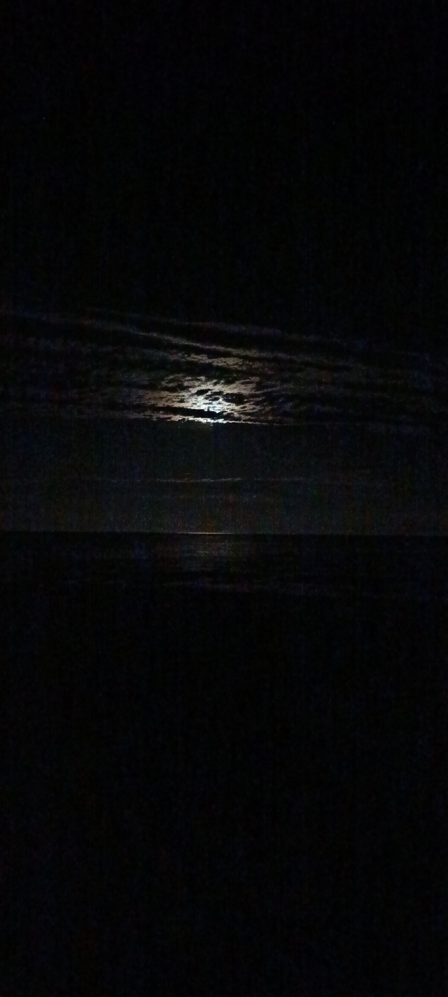 A nighttime, low-light photograph depicts a body of water reflecting moonlight. The scene is dark, with the water occupying the lower half of the frame and the sky above. The moon, partially obscured by clouds, casts a shimmering path across the water's surface. The clouds are dark and elongated, streaking horizontally across the sky. The overall impression is one of tranquility and mystery, with the limited light creating a stark contrast between the illuminated water and the surrounding darkness.