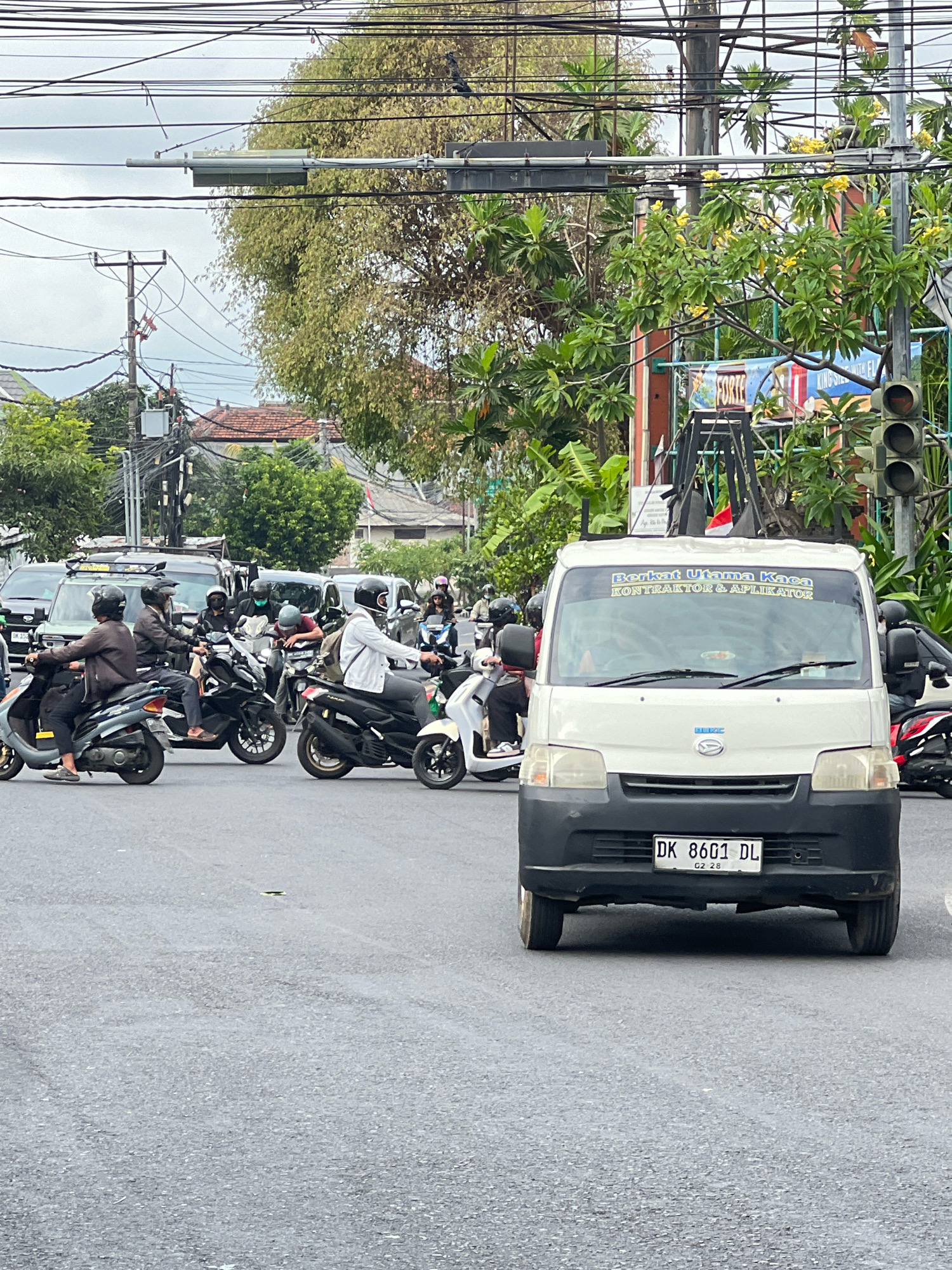 This image shows a street scene in Denpasar, Indonesia, likely at an intersection, characterized by heavy motorcycle traffic. A white van with the license plate DK 8601 DL is prominently featured in the foreground, its windshield displaying a yellow sticker with blue text that reads "BERKAT UTAMA KACA KONTRAKTOR & APLIKATOR". The van appears to be stopped, possibly in traffic. Numerous motorcycles, many with riders wearing helmets, are visible in the mid-ground, navigating the intersection. Some motorcycles are moving, while others appear to be waiting. The environment includes a traffic light, power lines, trees with green foliage, and buildings in the background, suggesting an urban setting. The road surface is asphalt, and the sky is overcast. The overall impression is one of a busy, everyday street scene in Denpasar.