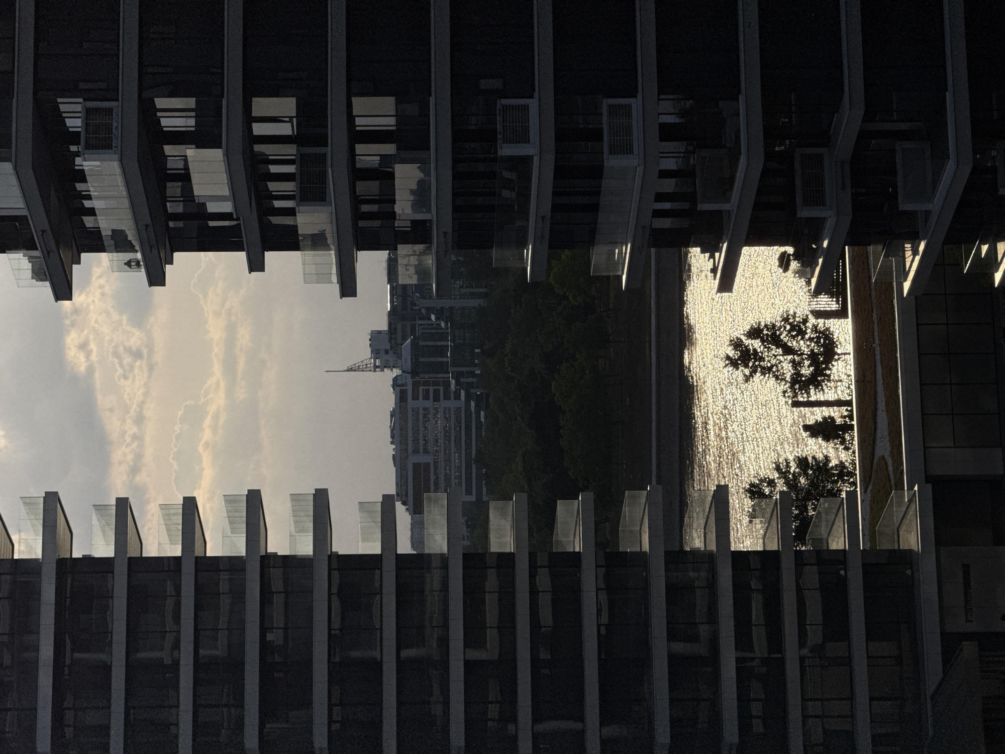 The image depicts a view of a cityscape seen through the structural elements of a building. The scene is vertically inverted. The top half of the image shows the tops of buildings and a sky with clouds, while the bottom half reveals a body of water with sunlight reflecting off the surface, creating a shimmering effect, and a tree silhouette. The framing building's architecture consists of dark vertical columns and balconies. The location is in an urban area with modern architecture.