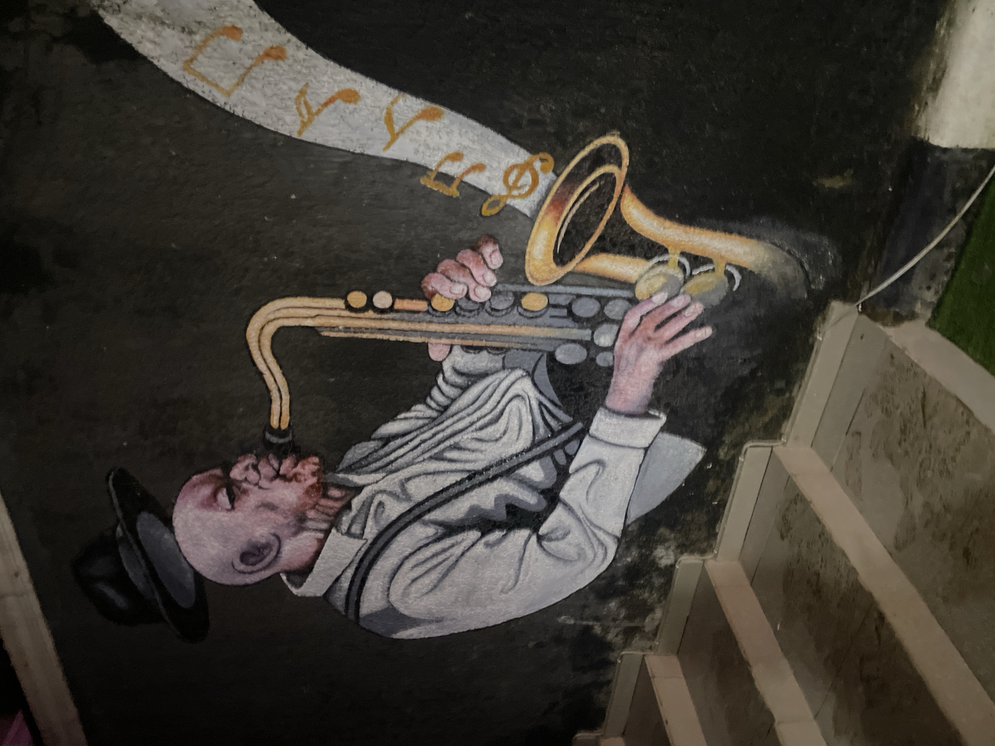 A mural depicts a bald Black man with a goatee playing a saxophone. He wears a black fedora, a white collared shirt, and suspenders. His body is angled to the left, and his hands are positioned on the saxophone keys. Musical notes in a light color with orange accents emanate from the bell of the instrument, moving towards the upper right of the frame. The background is dark. To the left of the mural are a series of concrete stairs. This mural is located on a wall in Guzape, Nigeria, adjacent to a staircase.