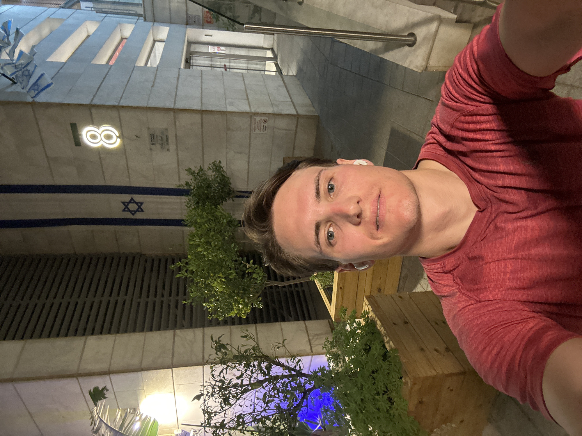 A selfie of a fair-skinned man with short brown hair and blue eyes, wearing a red t-shirt and a white earbud in his left ear. The man is in front of a building with a large Israeli flag and the logo of the Diamond Exchange District. The location is likely in Ramat Gan, Israel, based on the flag and logo. The environment includes urban elements such as trees, wooden structures, and walkways. The time is evening, as suggested by the lighting.