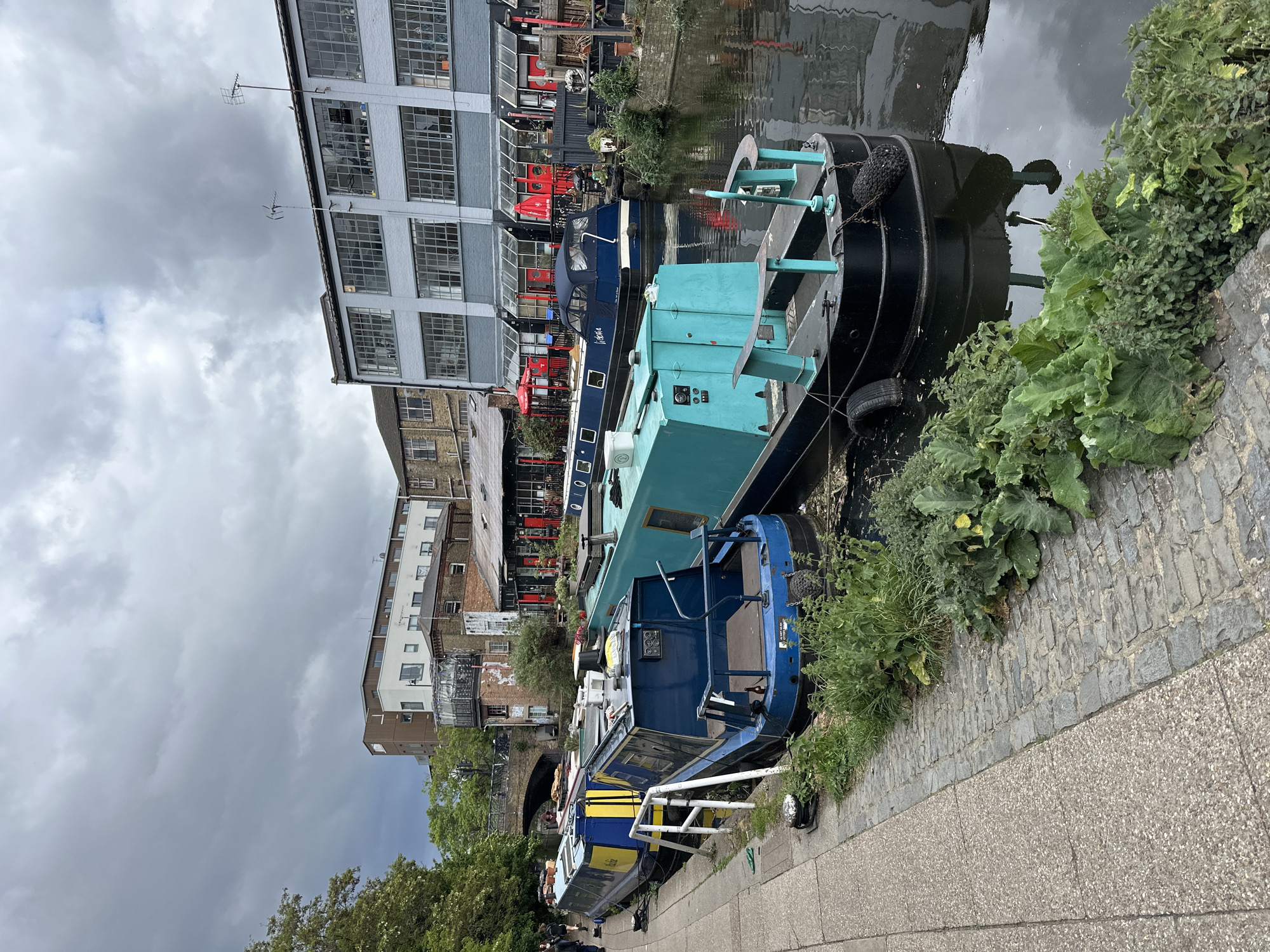 The image shows a section of the Regent's Canal in London, United Kingdom. Several canal boats are docked along the towpath, exhibiting various colors and designs, including black, teal, and blue. The boats are close to the edge, with the canal water visible. The towpath is paved with concrete and brick. Lush greenery and vegetation grow along the canal's edge. The background features buildings of mixed architecture, including a gray brick building with numerous windows and red accents and other residential or commercial structures. The sky is overcast with dark, cloudy skies overhead. The scene captures a typical waterside view of this part of London.
