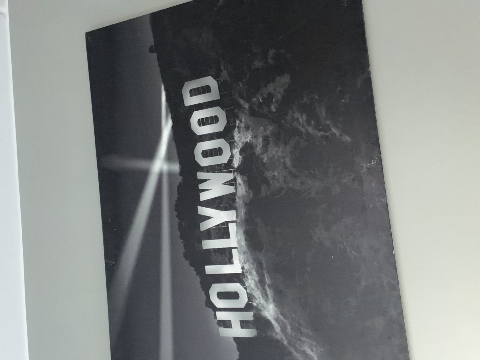 This black and white image displays a striking photograph or print of the iconic Hollywood sign. The sign, rendered in large, white, block capital letters spelling "HOLLYWOOD", is the central subject, appearing to be situated on a dark, textured hillside. The surrounding environment, both above and below the sign, exhibits a cloudy or rugged texture, adding to the dramatic atmosphere. Prominent streaks of bright light emanate from the left, cutting across parts of the sign, suggesting either a nighttime setting with spotlights or a stylized artistic effect. The entire image is rotated approximately 90 degrees counter-clockwise, orienting the "HOLLYWOOD" text vertically. No people or specific activities are visible. The image captures the grandeur and iconic status of the landmark. While the subject is the Hollywood sign located in California, USA, the media file itself is found in Cape Town, South Africa.