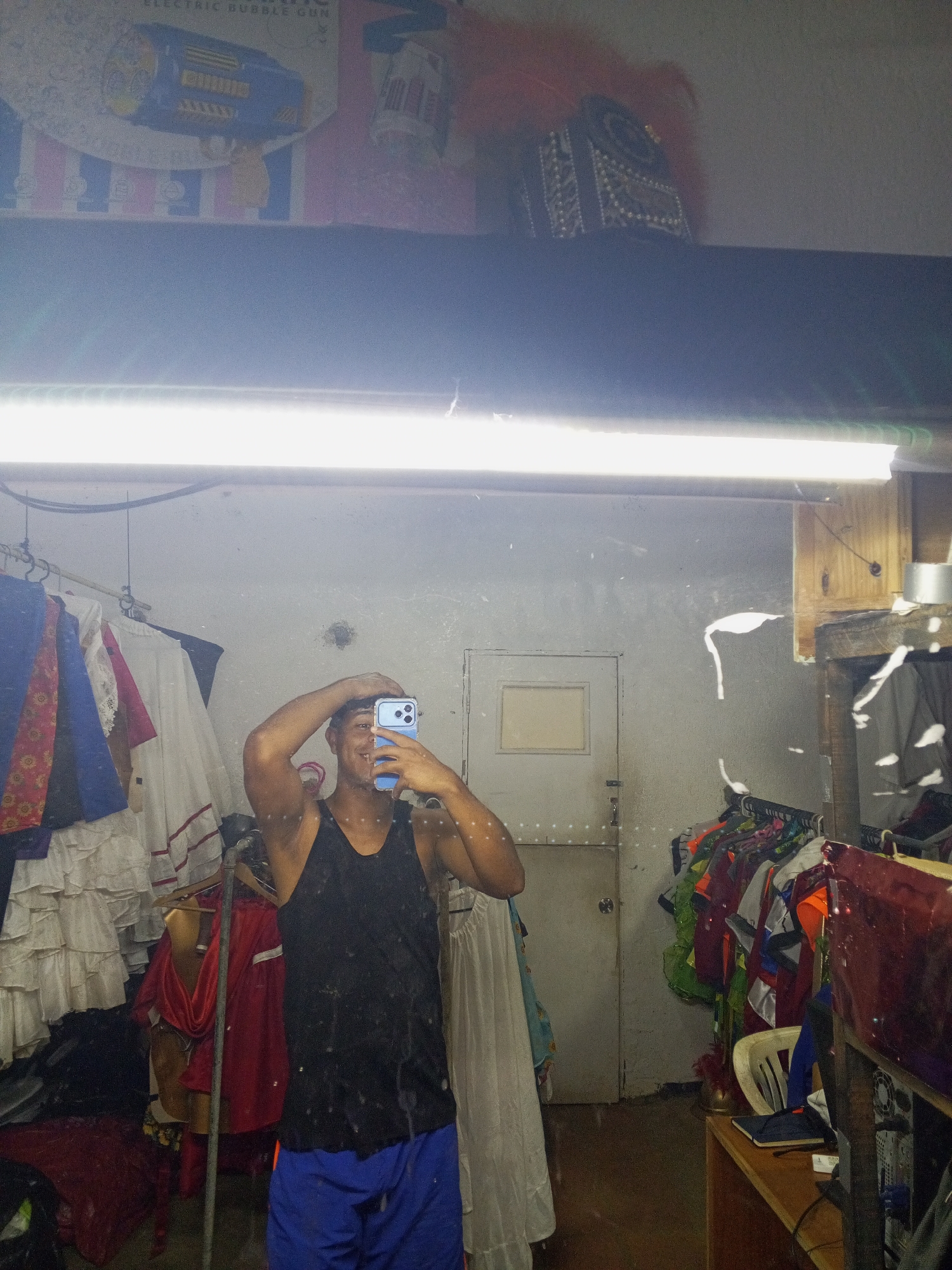 The image captures a mirror selfie taken indoors in what appears to be a cluttered room, possibly a storage area, dressing room, or workshop in Porlamar, Venezuela. The mirror itself is dusty or speckled.
A man with dark skin, wearing a black tank top and blue shorts, is the primary subject, reflected in the mirror. He is smiling slightly and holding a light blue smartphone to take the picture, with his other hand resting on his head.
Behind him, in the reflection, are various items. To the left, clothes, including white ruffled skirts and colorful garments, hang on racks. In the mid-background, a plain white door with a small rectangular window is visible. To the right, more clothes or costumes in bright colors like red, green, and orange are on hangers. A wooden shelf holds a white chair, a dark blue notebook, and part of a computer tower.
Above the mirror, resting on a shelf, are two distinct items: to the left, packaging for an "ELECTRIC BUBBLE GUN" showing a yellow and blue toy; to the right, an ornate, possibly decorative box with gold and black patterns, accompanied by red feathery material.
The room is illuminated by a bright fluorescent light fixture, which reflects prominently across the top of the mirror. The overall impression is one of a casual, lived-in, and somewhat untidy space.