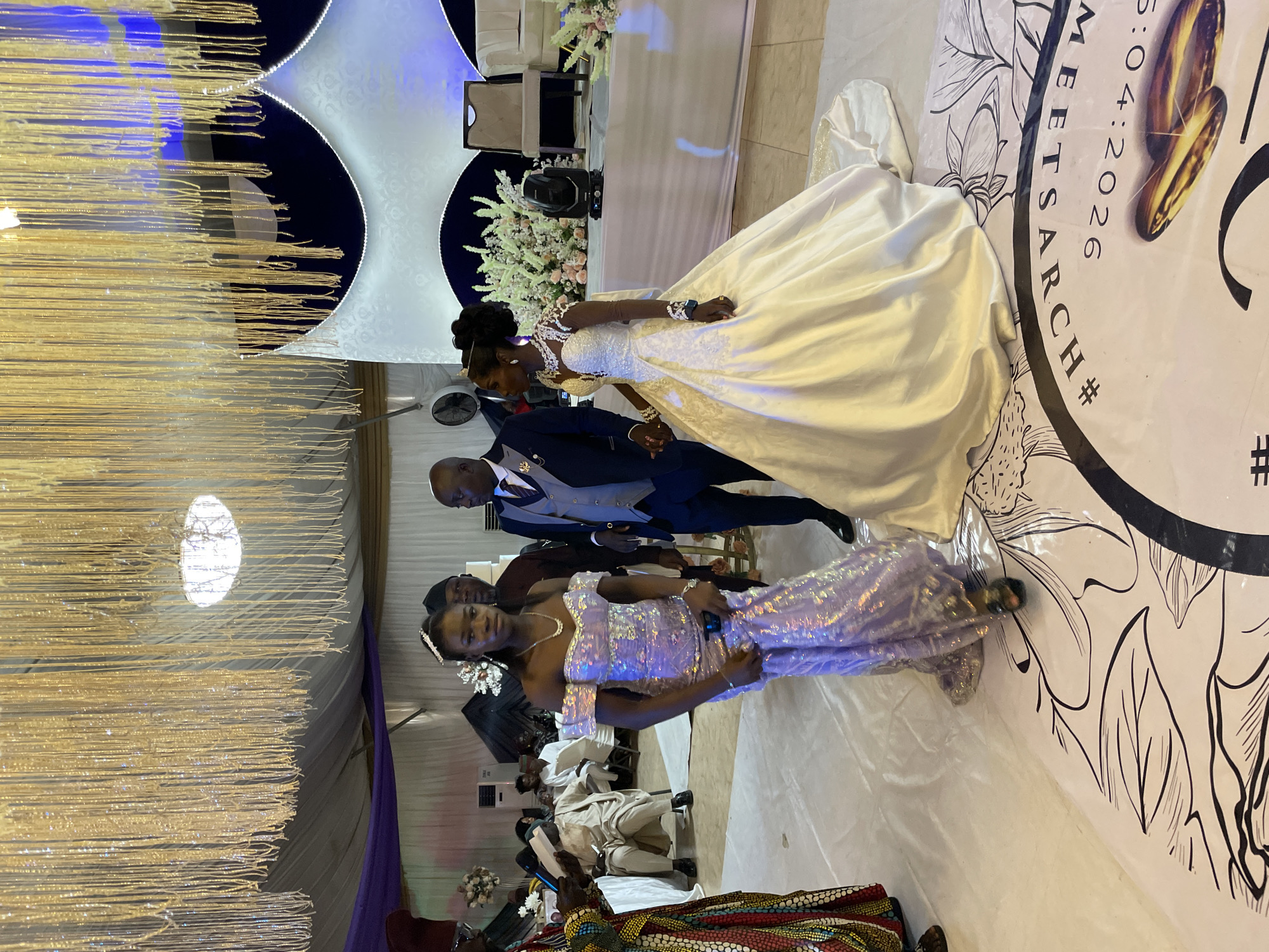 This image captures a vibrant indoor wedding reception in Jos, Nigeria, characterized by elaborate decor and formal attire. The scene is well-lit with artificial lighting, suggesting an evening event.
In the mid-ground, the bride and groom are featured prominently, holding hands. The bride is dressed in a classic white wedding gown with detailed lace on the bodice and long sleeves, her hair styled elegantly. The groom wears a dark suit, a lighter waistcoat, and a tie. They stand near a decorated table adorned with white and light pink floral arrangements.
In the foreground, a woman in a shimmering, off-the-shoulder lavender/lilac gown is seen holding a microphone, suggesting she is either a performer, an event host, or delivering a speech. She has a hair accessory resembling a small tiara.
The venue is richly decorated with hanging golden, string-like elements from the ceiling that sparkle under the lights. Blue and white fabric panels form an elegant backdrop. The floor features a large, white circular decal or patterned carpet with black illustrative designs. Visible text on this floor design reads "MEETS ARCH" and "5:04:2026", accompanied by a hashtag symbol, indicating a wedding date and possibly a couple's or event's hashtag. Two golden wedding rings are also depicted on the floor, reinforcing the celebratory marriage theme. Guests are visible in the background, seated and dressed in formal wear. The overall mood appears joyful and festive.