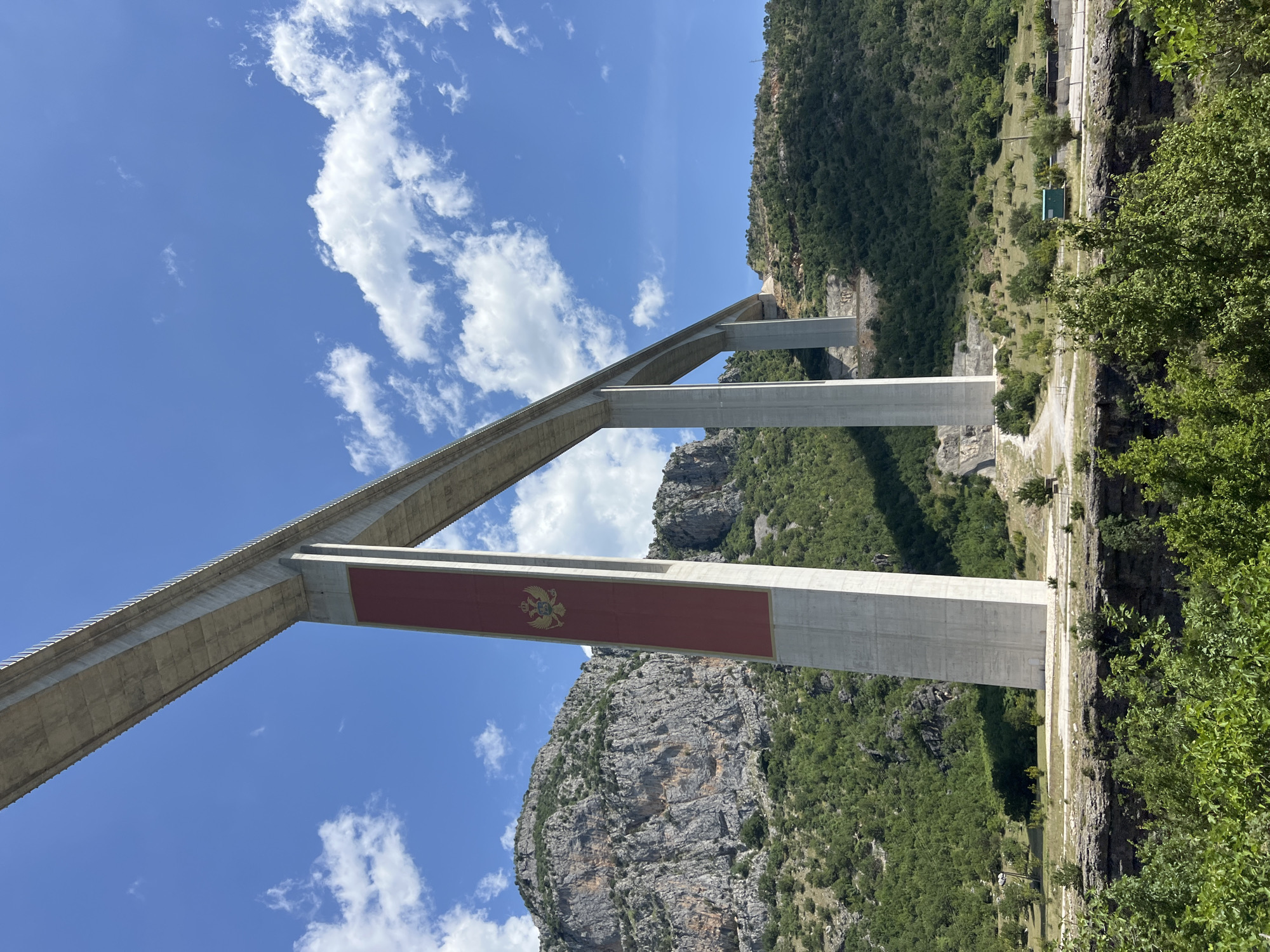 The image captures a view of the Moračica Bridge, a landmark in Bioče, Montenegro. The bridge's concrete support structure is visible against a backdrop of a blue sky with scattered white clouds and mountainous terrain covered in dense green vegetation. A large red panel adorned with a golden emblem, likely the coat of arms of Montenegro, is positioned on one of the bridge's support pillars. The surrounding environment includes rocky cliffs and a road running alongside the bridge's base.