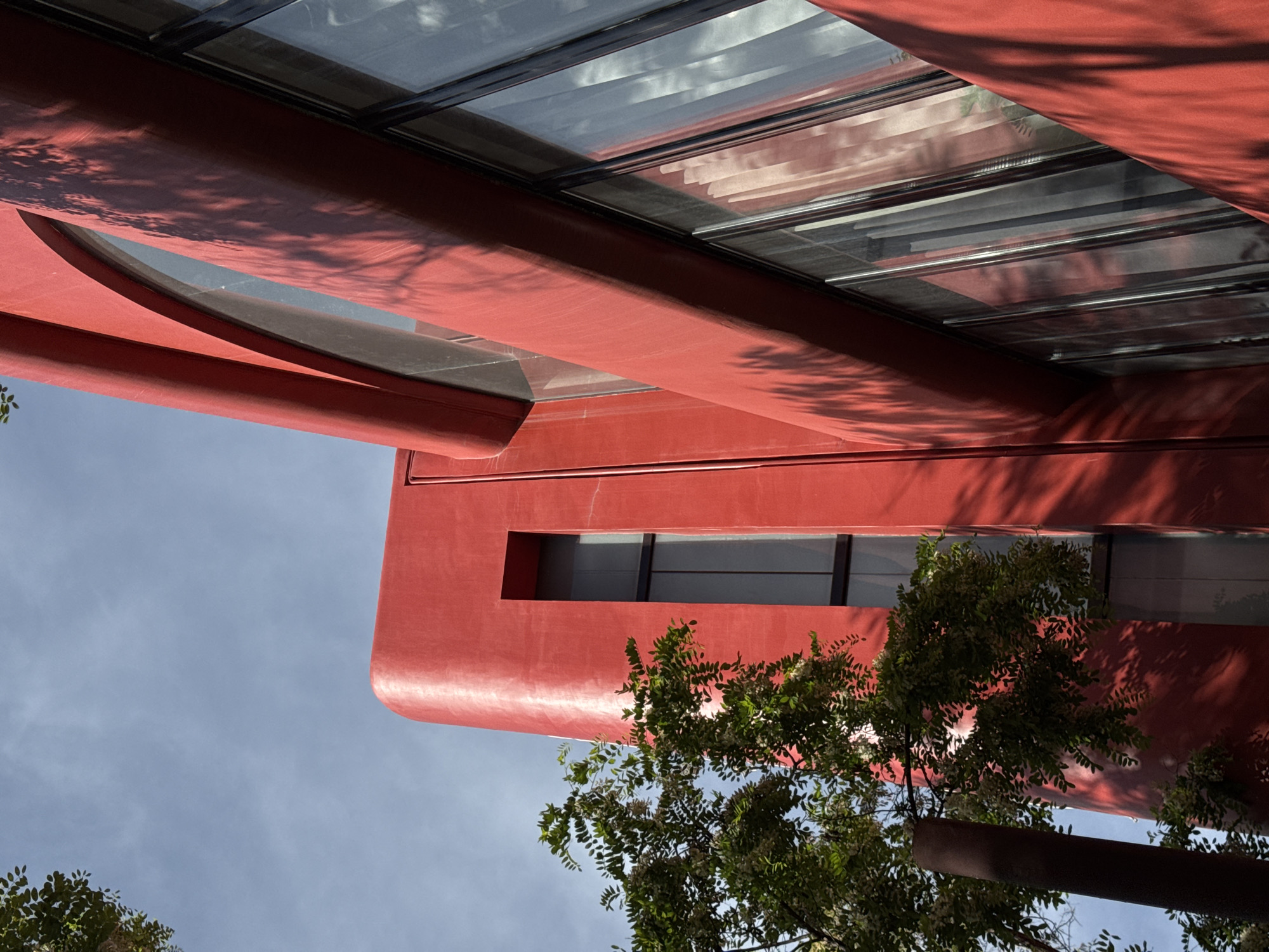 The image depicts a low-angle view of a modern, red-painted building against a partly cloudy sky. The building features rounded architectural elements, including a large rounded window and straight-edged windows. The surface of the building is textured with shadows, likely cast by nearby trees, one of which is visible at the bottom of the frame. The building has black-framed windows. The image location is Yerevan, Armenia.