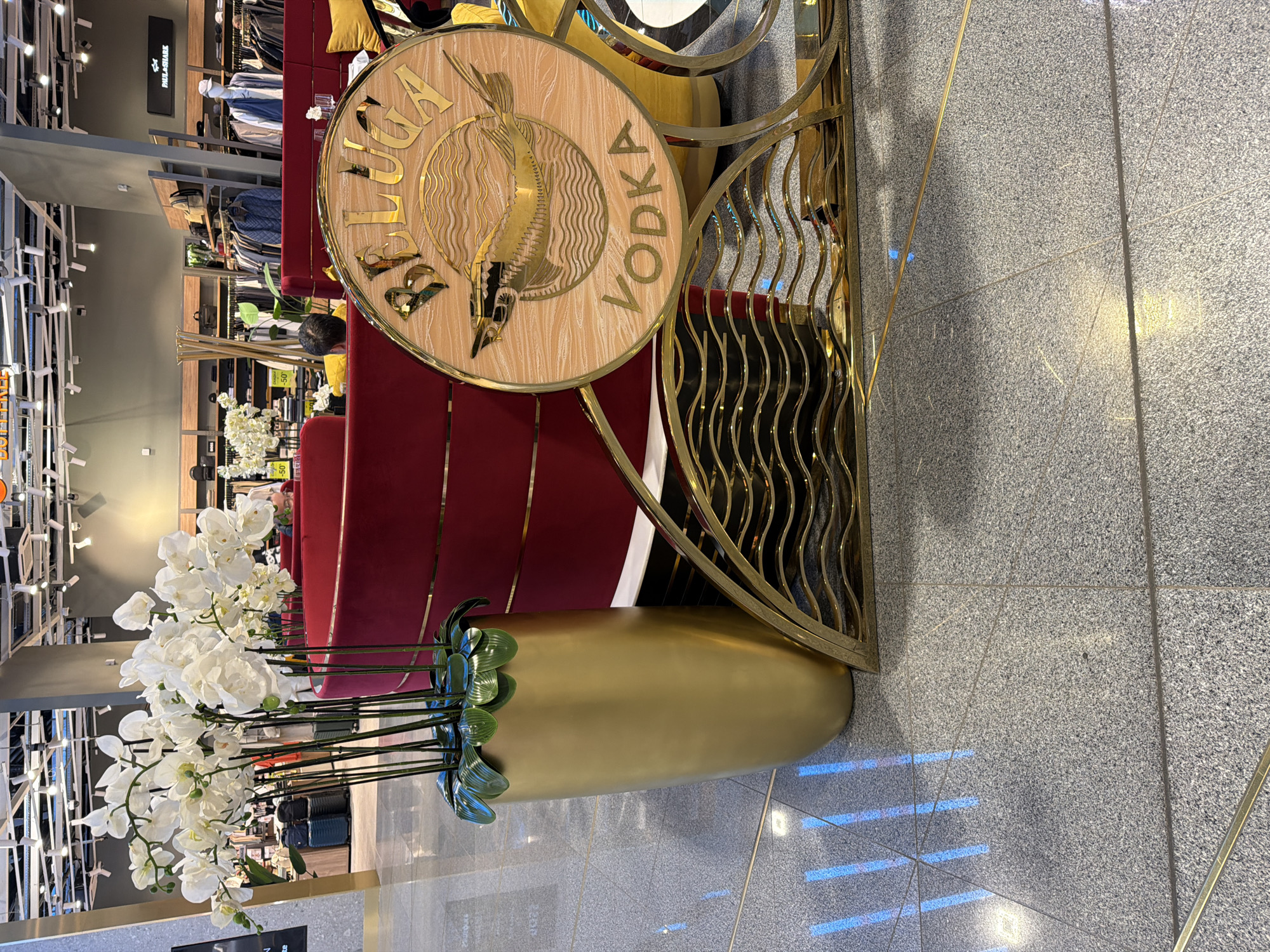 The image shows a display of Beluga Vodka in what appears to be a retail setting, possibly a shopping mall or a high-end store, located in Unknown, Unknown. The display features a large, circular sign made of light-colored wood with a gold-colored metal trim. On the sign, the words "BELUGA" and "VODKA" are written in large, gold, three-dimensional letters, flanking an image of a sturgeon. The sign is part of an ornate gold-colored metal structure with a wavy design that serves as a backdrop to a red velvet couch or seating area. In front of the display stands a tall, gold-colored vase containing white orchids, adding an elegant touch. The floor is made of light gray tiles with darker flecks, and the lighting is bright, indicating an indoor environment. The background shows shelves stocked with clothing, suggesting the location is inside a department store.