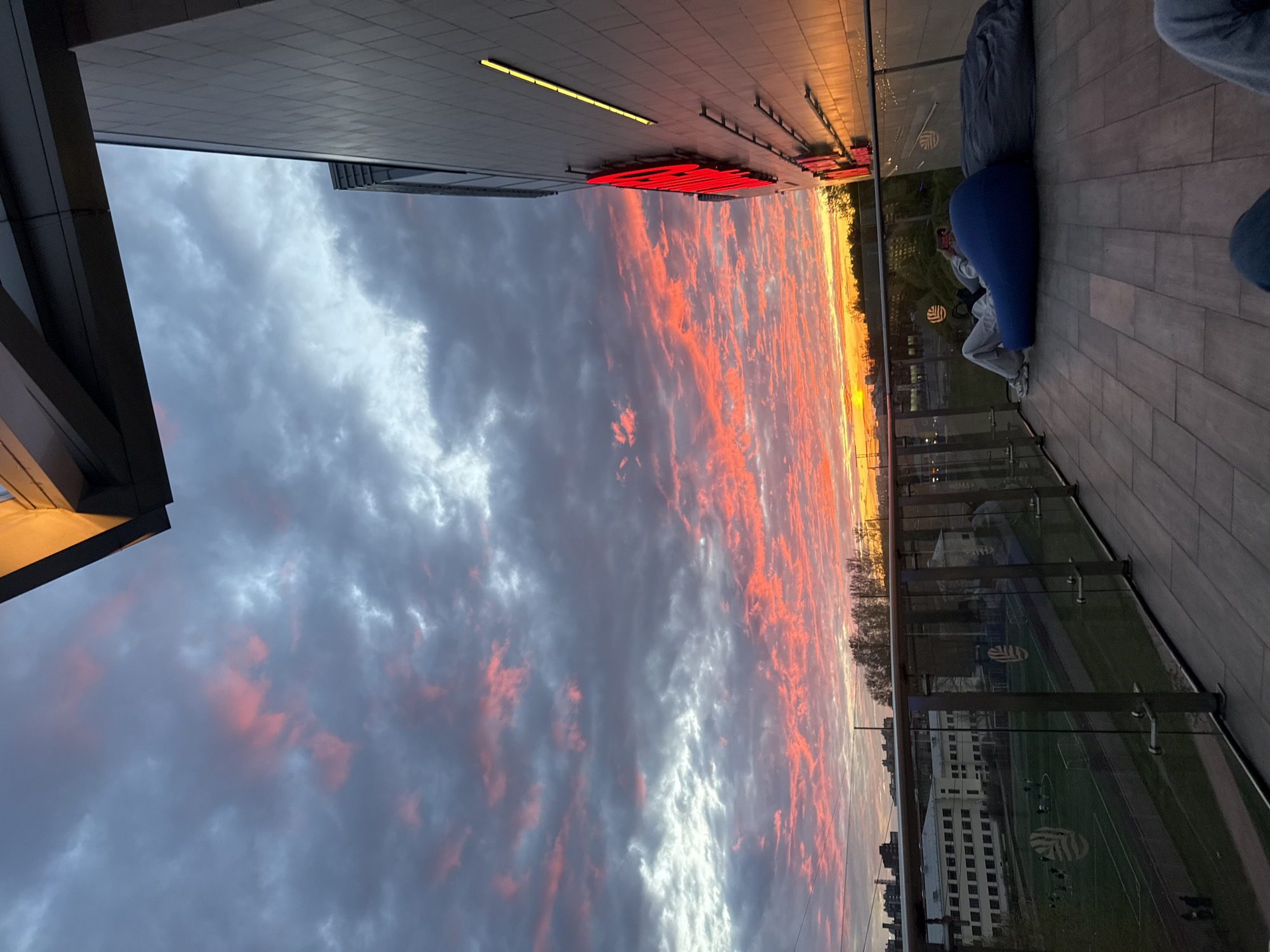 The photograph depicts a cityscape, likely taken in Saint Petersburg, Russia, given the architecture and urban layout. The sky is dominated by a dramatic sunset with dark gray clouds streaked with vibrant pink and orange hues. The view is partially framed by a modern building with a gray façade and a prominent red neon sign.
To the right of the frame, a section of an elevated outdoor space is visible, featuring a wooden floor, glass railing, and several beanbag chairs. A person, possibly taking a photograph, is seated on one of the beanbag chairs. Behind the railing, a glimpse of a sports field and buildings below can be seen.