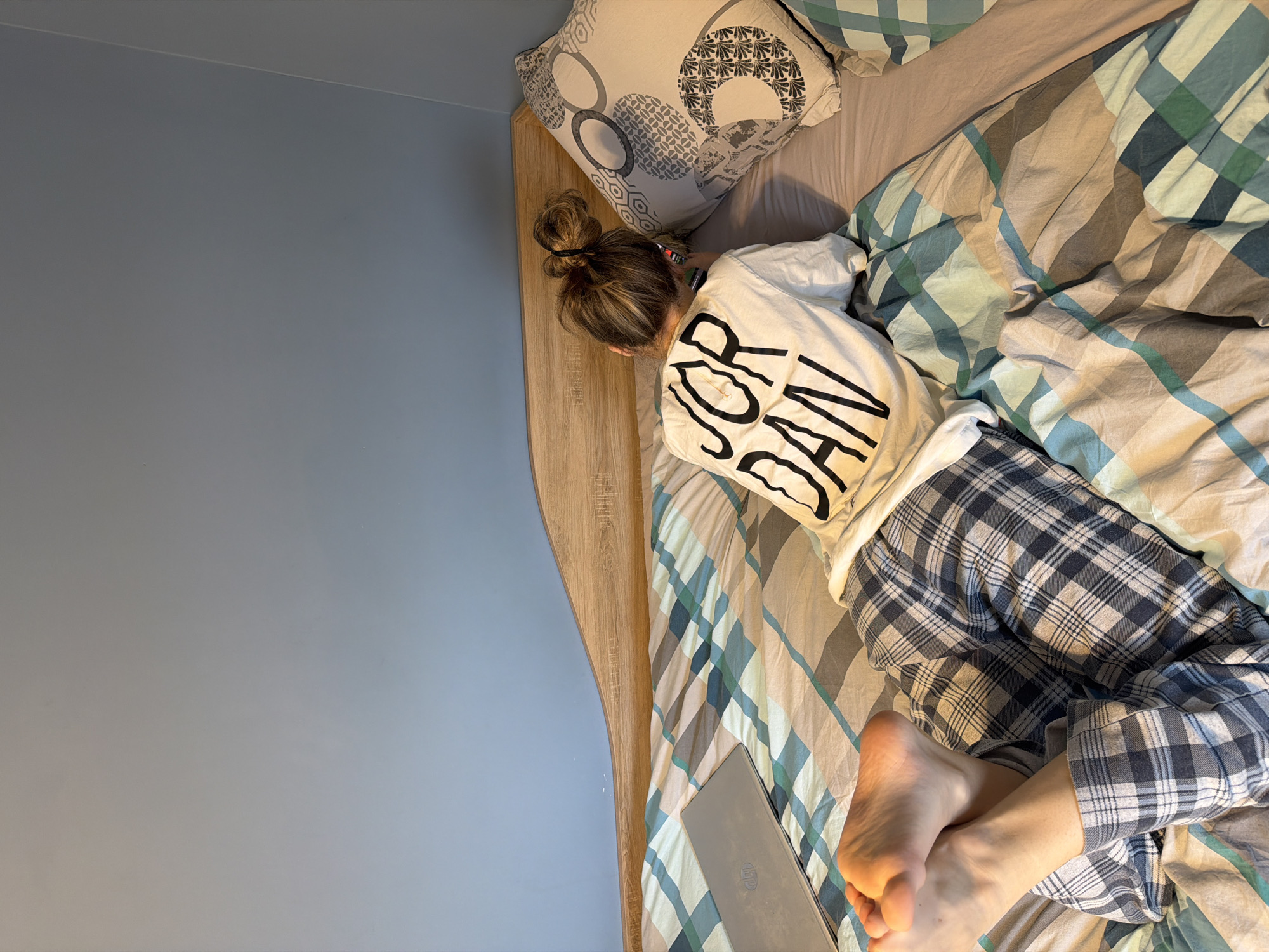A person is lying on a bed in a room with light blue walls and a light wood headboard. The person has their hair in a bun and is wearing a white t-shirt with "JORDAN" printed on the back, and plaid pajamas. The bed has a patterned pillow and a striped comforter with shades of brown, green, and blue. A laptop is visible at the foot of the bed. No information indicates a specific location in the city of Plovdiv, Bulgaria.