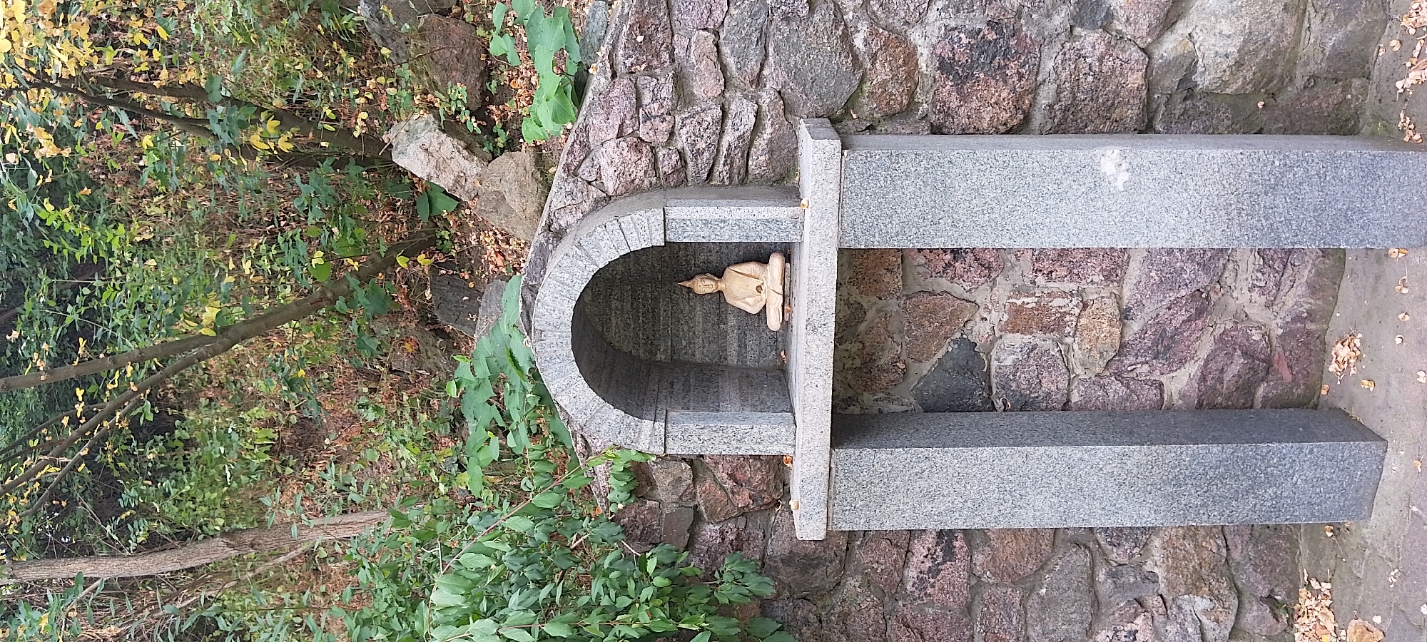 A small, light-colored statue of the Buddha in a meditative pose sits within a grey granite niche, which is part of a larger grey granite structure. This structure is mounted on a rough stone wall. The niche has an arched opening. The scene is set outdoors, with lush green foliage and trees forming the background. The ground in front of the structure is paved, and scattered dry leaves suggest an autumn season. The image appears to be taken in the city of Cherkasy, Ukraine.