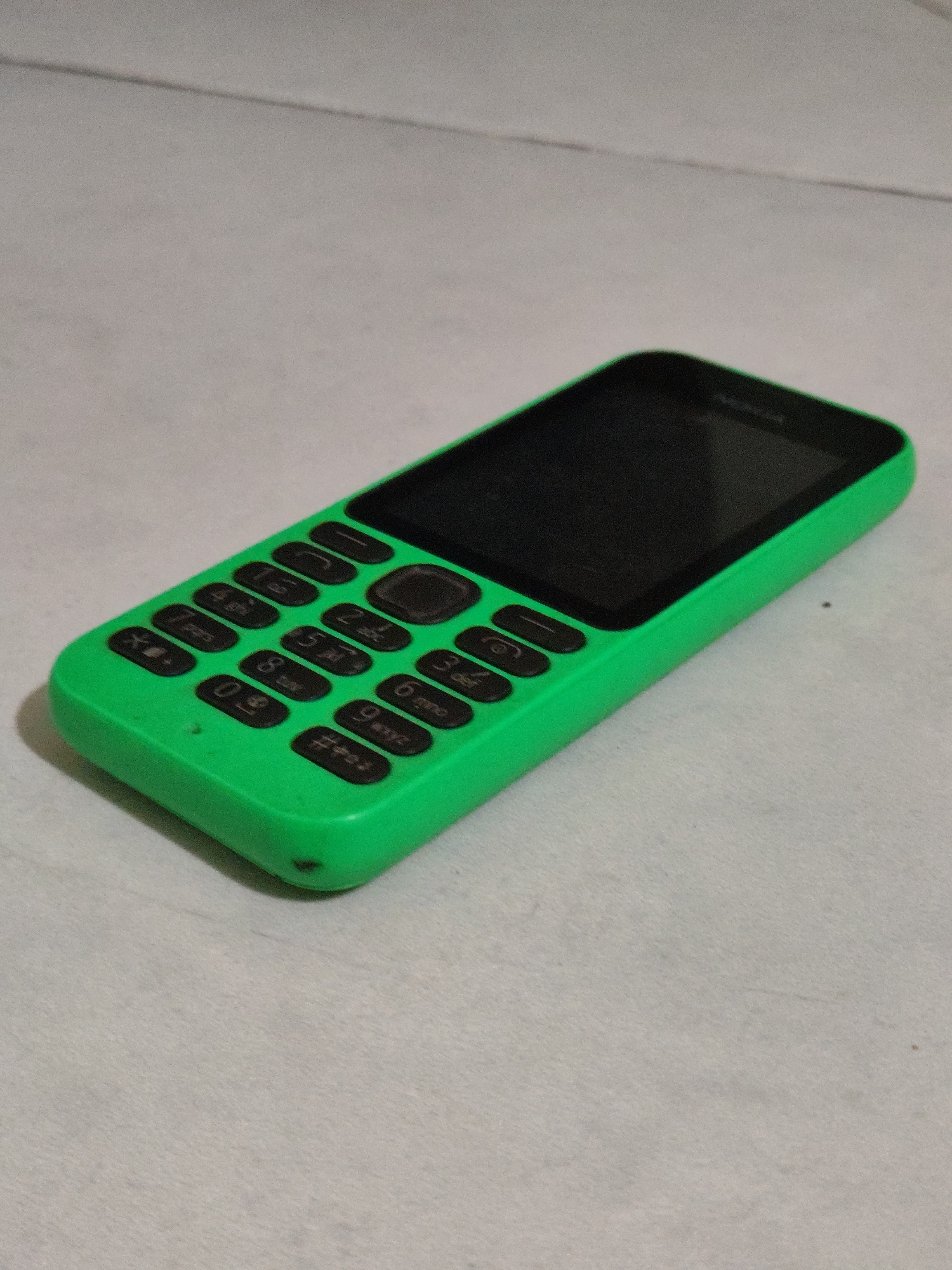 A bright green Nokia feature phone with a black screen and keypad lies on a textured, light gray surface. The phone is positioned at an angle, with the top left corner closer to the viewer. The keypad has standard numeric buttons (0-9), along with asterisk and hash symbols. Above the keypad is a navigation button cluster. The screen area is dark, suggesting it is off or displaying a black image. The overall impression is of a simple, functional mobile device resting on a neutral background. The location of Bulakrejo, Indonesia is not discernible from the image itself.