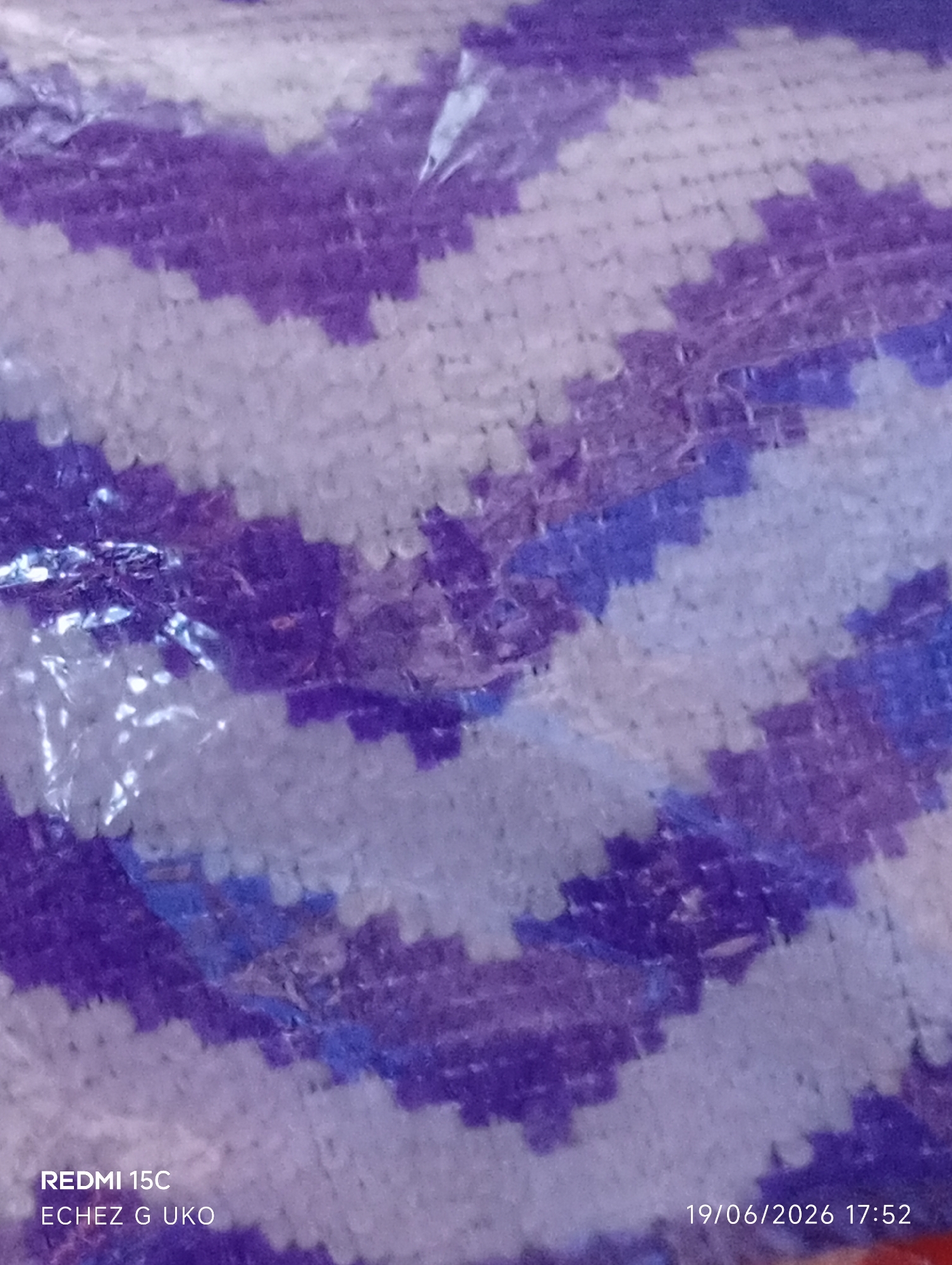 The image shows a close-up, blurred view of a textured fabric with a chevron pattern. The colors are primarily shades of purple, white, and a lighter blue, with some areas appearing more saturated than others due to light reflections on what seems to be a plastic covering. The fabric appears to be a knitted or crocheted material. In the bottom left corner, the text "REDMI 15C ECHEZ G UKO" is visible, indicating the phone model used for the photo and potentially a person's name or a location. In the bottom right corner, a date and time stamp reads "19/06/2026 17:52". There are no discernible people, actions, or interactions. The environment is not specified beyond the visual details of the fabric. The location in Jalingo, Nigeria, is not visually represented in the image itself, but is provided as contextual information.