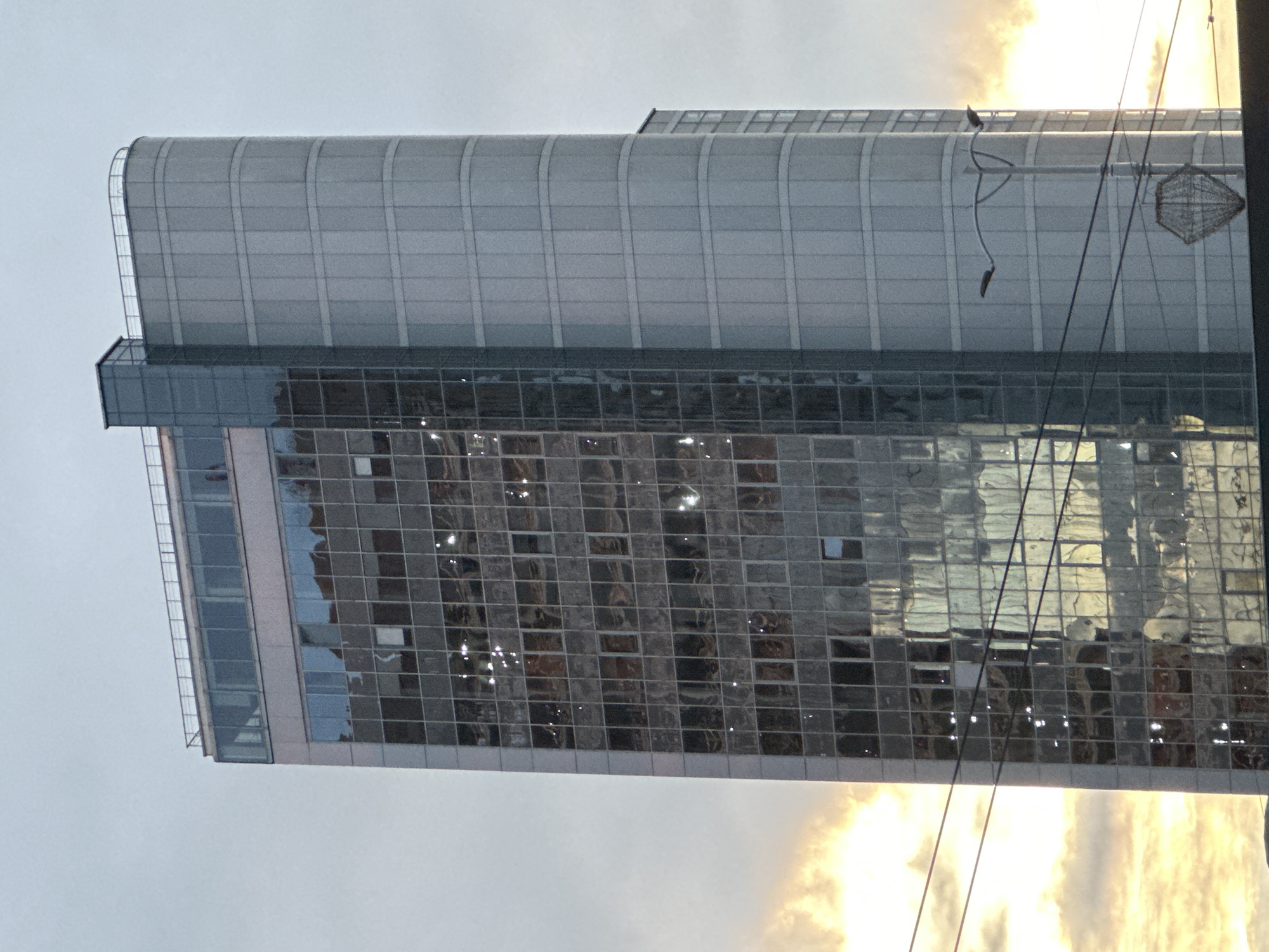 This image depicts the Prime Business Center building in Minsk, Belarus, located at Niamiha Street, 5. The tall, modern structure dominates the frame, viewed from a slightly low angle.
The building features two distinct architectural styles: the left side presents a flat, multi-story glass facade composed of numerous square and rectangular panels, reflecting an overcast sky with warm, golden light from the lower left, indicating either sunrise or sunset. Several reflections appear as bright, indistinct glares or distorted patterns. The right side of the building is a curved, non-reflective surface, clad in horizontal segments of light grey paneling. A vertical, dark grey, box-like structure, possibly an elevator shaft or service core, connects the two main sections near the top. The rooftop includes a smaller, recessed glass enclosure and a metal railing spanning across both architectural forms.
The sky in the upper right is a muted, light grey, while the lower left is illuminated by a strong, warm, yellowish-orange glow, consistent with atmospheric light at dawn or dusk, reflecting off the glass facade. In the lower foreground, multiple dark electrical or communication wires traverse horizontally across the scene. A dark grey street lamp with two curved light fixtures extends from the lower right. Below the street lamp and wires, a white, open-frame, diamond-shaped decorative object is suspended, possibly a light fixture. No discernible human figures or explicit actions are present within the scene.