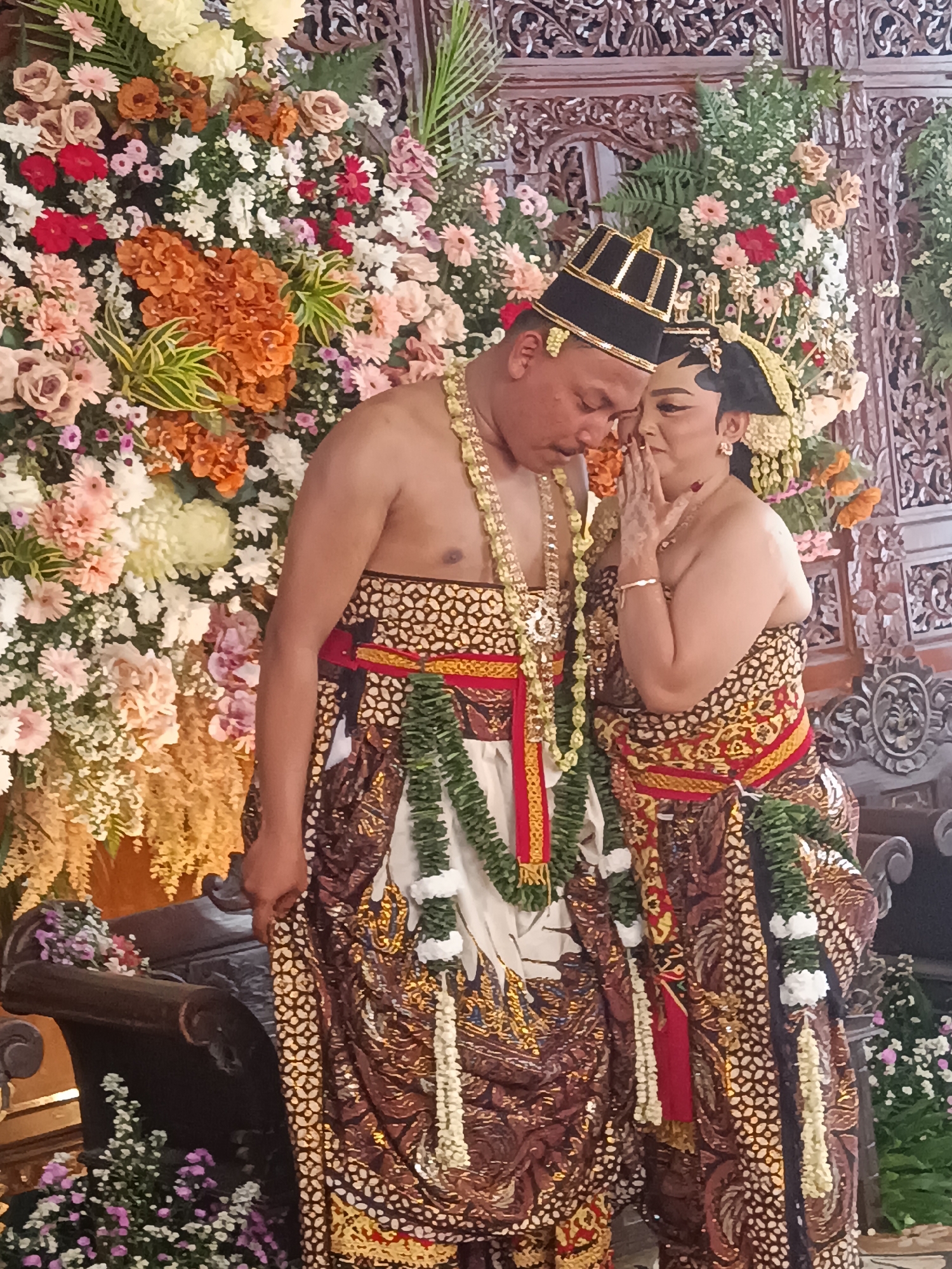 This image captures a traditional Indonesian wedding ceremony, likely in Depok, Indonesia, given the attire and decorations. A couple, dressed in ornate, traditional Javanese wedding attire, is the central focus. The groom wears a black and gold headdress, a patterned sarong, and is adorned with multiple beaded necklaces and a floral garland. He is leaning in towards the bride, with his eyes closed. The bride, wearing a matching headdress and a richly patterned sarong, has her hand cupped near her mouth and is looking at the groom with a tender expression. The background is a lavish display of flowers, primarily in shades of pink, orange, white, and red, with lush greenery and intricately carved wooden panels adding to the elaborate decor. The overall scene conveys a sense of intimacy and cultural richness associated with traditional Indonesian weddings.
