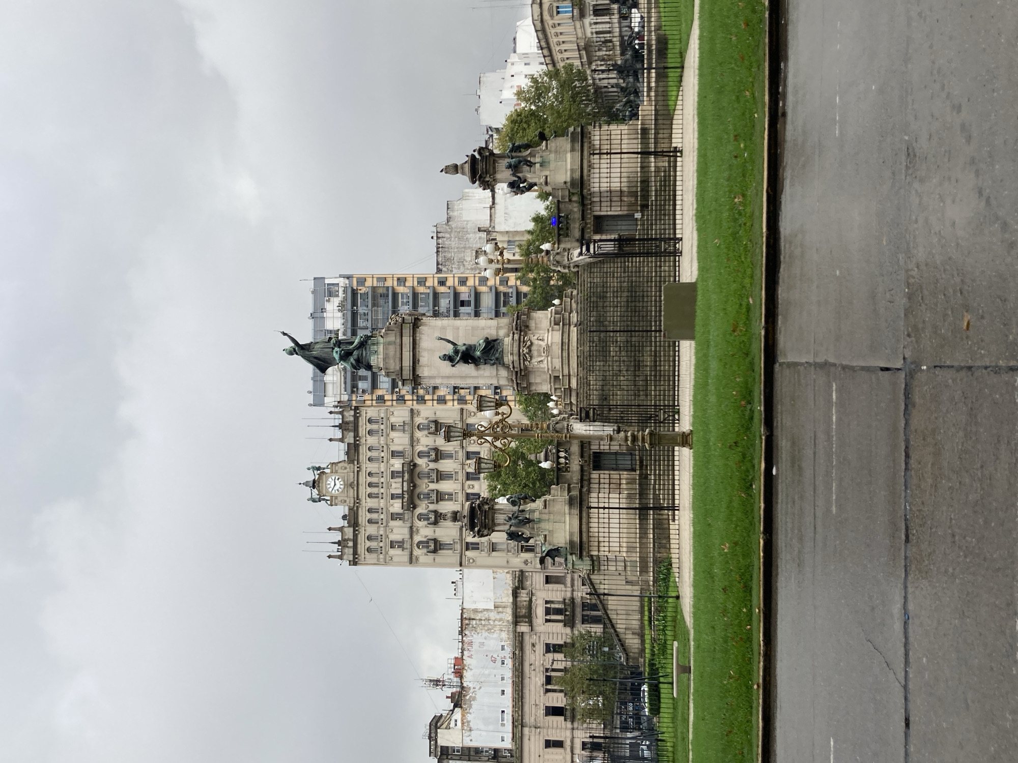 The image, oriented correctly, depicts a grand urban scene under an overcast sky. In the foreground, a wet asphalt road stretches horizontally, with a strip of vibrant green grass and a black metal fence running parallel to it on the left. The wetness of the road and pavement suggests recent rain or damp conditions.
Beyond the fence and grass lies a large, ornate monument, likely a historical landmark, featuring multiple dark bronze statues atop a stone base. One prominent statue depicts a figure with an outstretched arm, pointing upwards. The monument is intricately designed with classical elements and what appears to be decorative lampposts or light fixtures.
Behind the monument, a collection of buildings forms the backdrop. The most prominent building directly behind the monument is a multi-story structure with an older, classical architectural style, characterized by numerous arched windows and decorative cornices. It features a clock tower on its upper section, though the exact time is not clearly visible. Other buildings in the distance appear to be a mix of older and more modern structures, some showing signs of wear or renovation. Air conditioning units are visible on the facades of some buildings, blending modern elements with the older architecture.
The sky is a uniform, light grey, heavily overcast, indicating a cloudy day with diffuse lighting. No specific human activity is visible, making the scene feel quiet and contemplative. The overall impression is that of a historic city square or a grand avenue. Visible text is minimal and difficult to discern clearly; some faint numbers like "1247" and "1280" are barely legible on a building in the background.