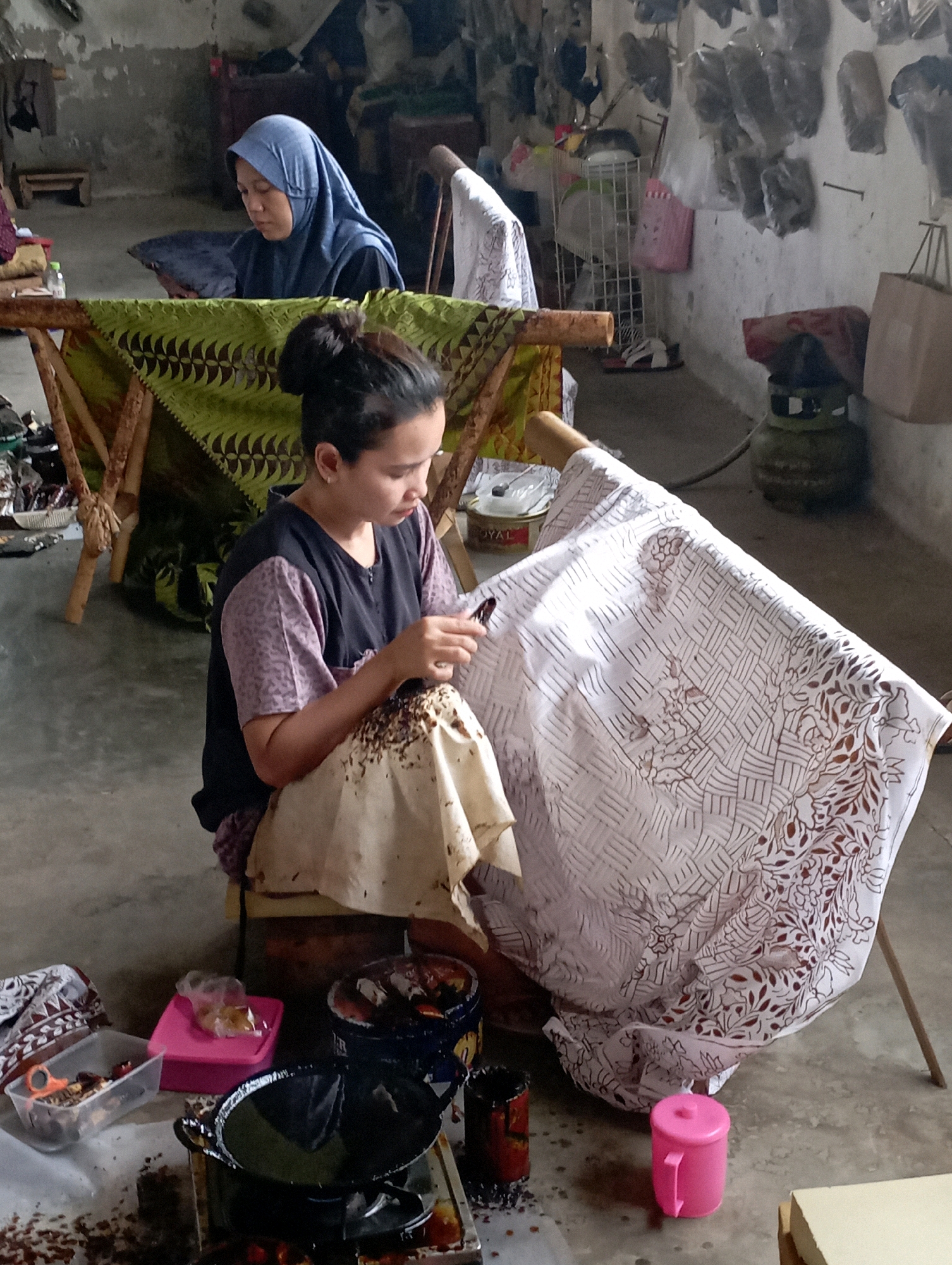 In a dimly lit workshop in Tasikmalaya, Indonesia, two women are engaged in the art of batik. The woman in the foreground, with her hair pulled back in a bun, is seated on a low stool and meticulously applying wax to a large piece of white fabric using a canting tool. The fabric, draped over a wooden frame, features intricate patterns of geometric shapes and floral motifs. Her attire consists of a purple and black patterned blouse. Behind her, another woman, wearing a blue headscarf, is seated at a table, also appearing to be involved in the batik process. The workshop environment is rustic, with concrete floors and walls that show signs of age. Various batik-making supplies and tools are scattered around, including a wok-like pan on a portable stove in the foreground, suggesting the use of hot wax. Shelves and racks in the background hold other batik fabrics and materials. The scene conveys a sense of focused craftsmanship and traditional artistry.