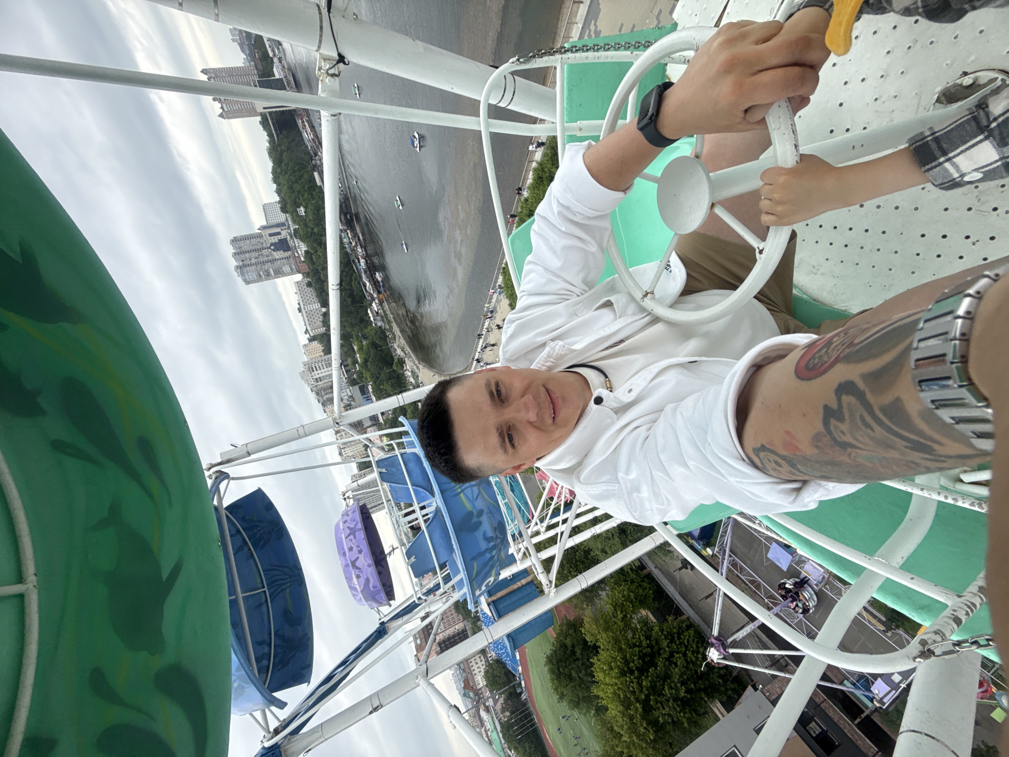 A man and a child are on a Ferris wheel in Vladivostok, Russia. The man is in the foreground, holding a white steering wheel. He is wearing a white shirt and khaki shorts. The child is to his right, also holding the steering wheel. The Ferris wheel is white with green accents. In the background, there is a river with boats and buildings along the shore. The sky is cloudy. The Ferris wheel is located at the waterfront park in Vladivostok, near the Sports Harbor. The Ferris wheel offers panoramic views of the city, the Amur Bay, and the Eastern Bosphorus Strait. The architecture of the buildings in the background suggests a mixed style, common in many Russian cities. The presence of boats on the water indicates an active port and recreational area.