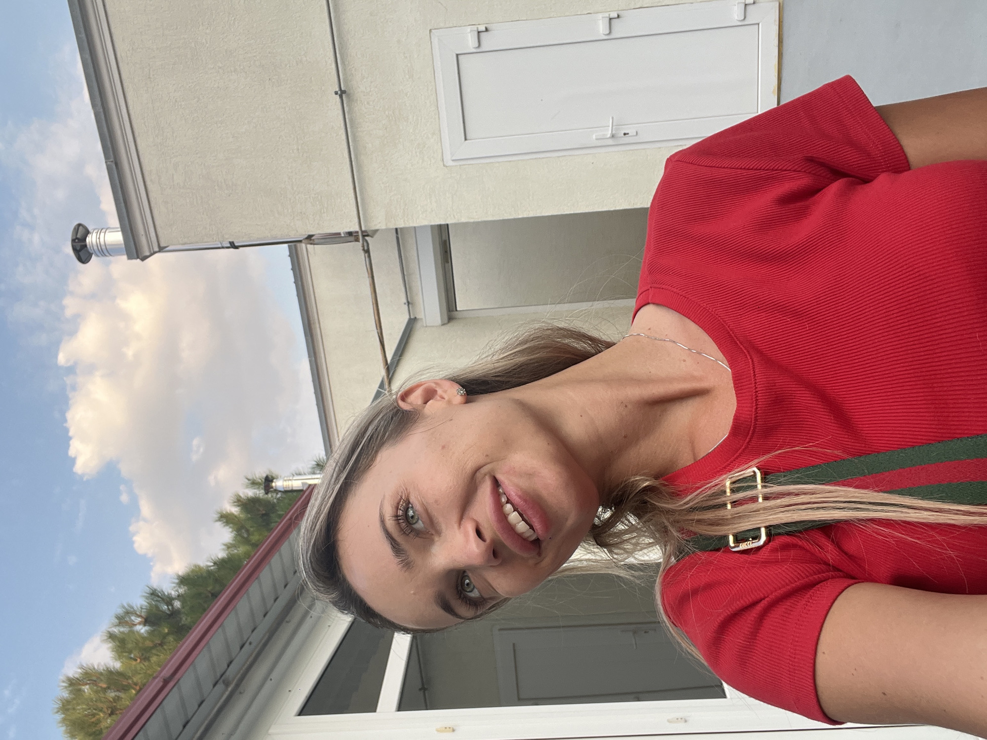 In Fontanka, Ukraine, a fair-skinned woman with long blonde hair is captured in a selfie. She is wearing a red, ribbed t-shirt with a green and red striped strap. Her head is tilted slightly upwards, and she is looking to her left with a gentle smile. The background features a pale yellow building with a white door and a metal chimney on the roof. To the left of the building, a portion of a red-roofed structure and green trees are visible under a sky with scattered clouds. The lighting suggests it is daytime.