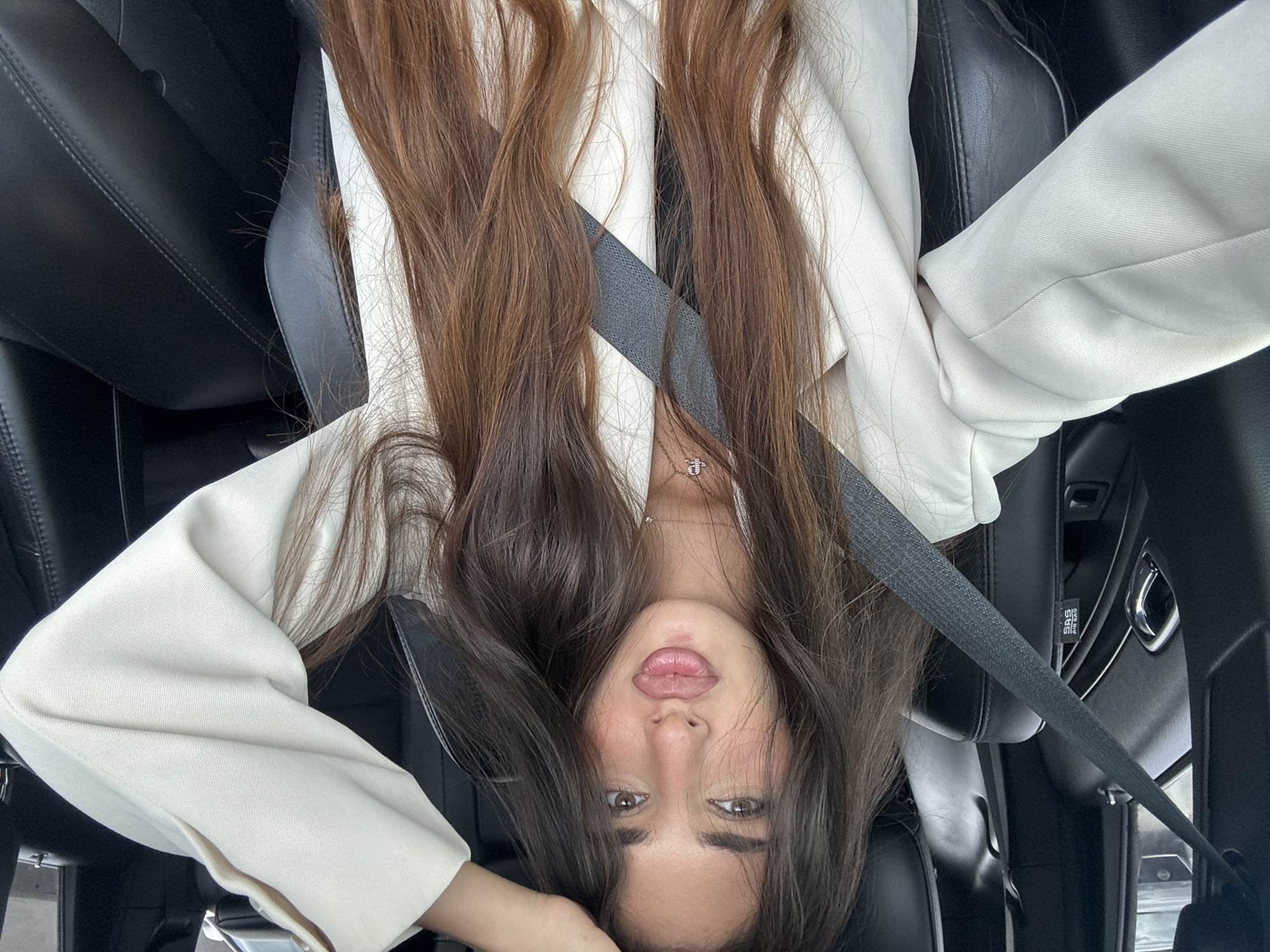 The image shows a young woman with long, light brown hair wearing a white blazer. She is in a car, wearing a seatbelt. The interior of the car has black leather seats. The image was taken indoors. It is not possible to verify the location of North Miami Beach, United States of America.