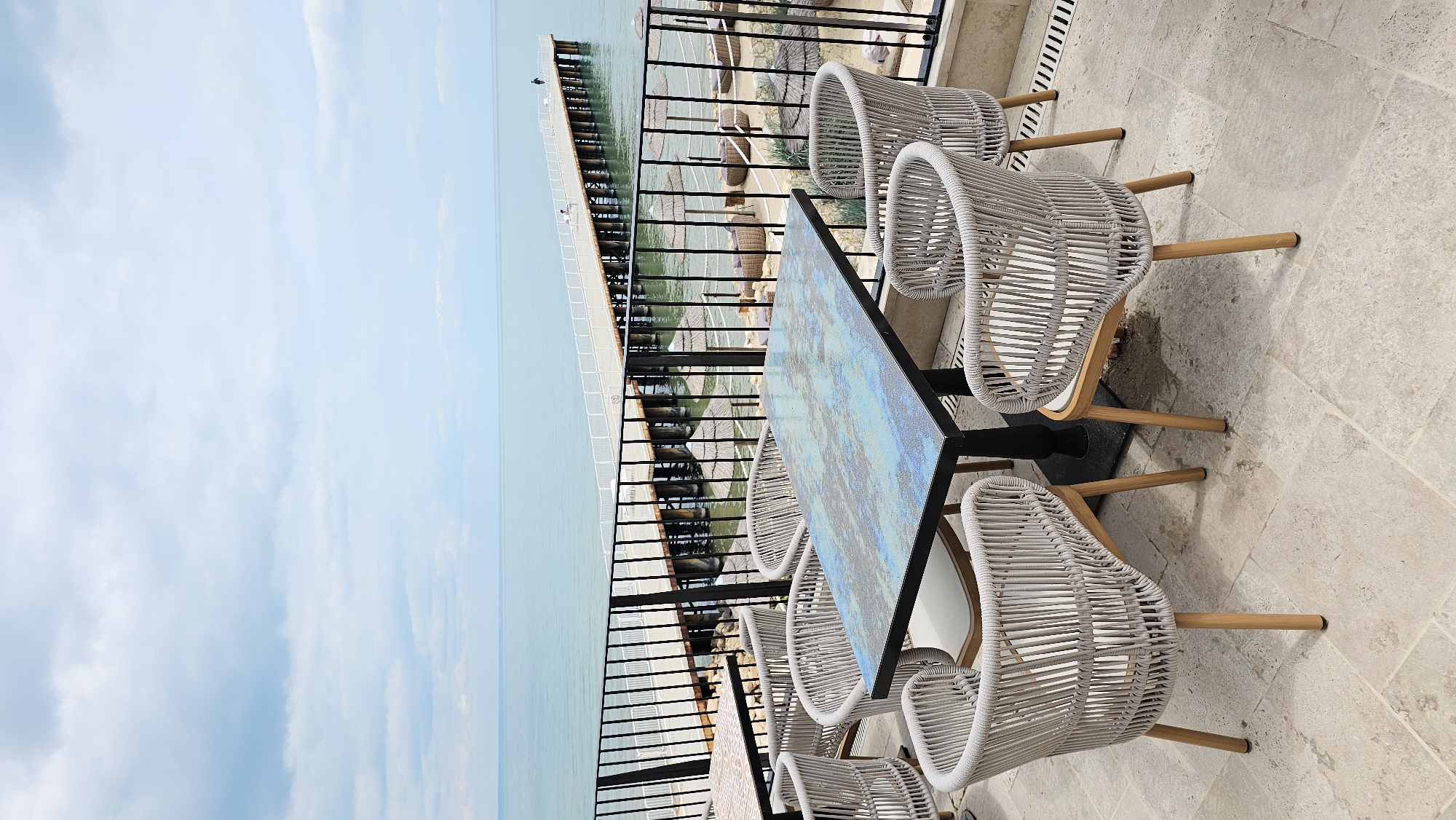 The scene depicts an outdoor dining area, seemingly on a balcony, overlooking a seascape, likely in Varna, Bulgaria. There is a table surrounded by several white wicker chairs. The table has a multicolored, patterned surface with a black frame and base. Beyond the balcony railing, there is a long pier or structure extending into the water. The sky is clear and pale blue, suggesting a sunny day. The floor of the dining area is stone. The overall atmosphere suggests a relaxed, upscale dining experience with a waterfront view.