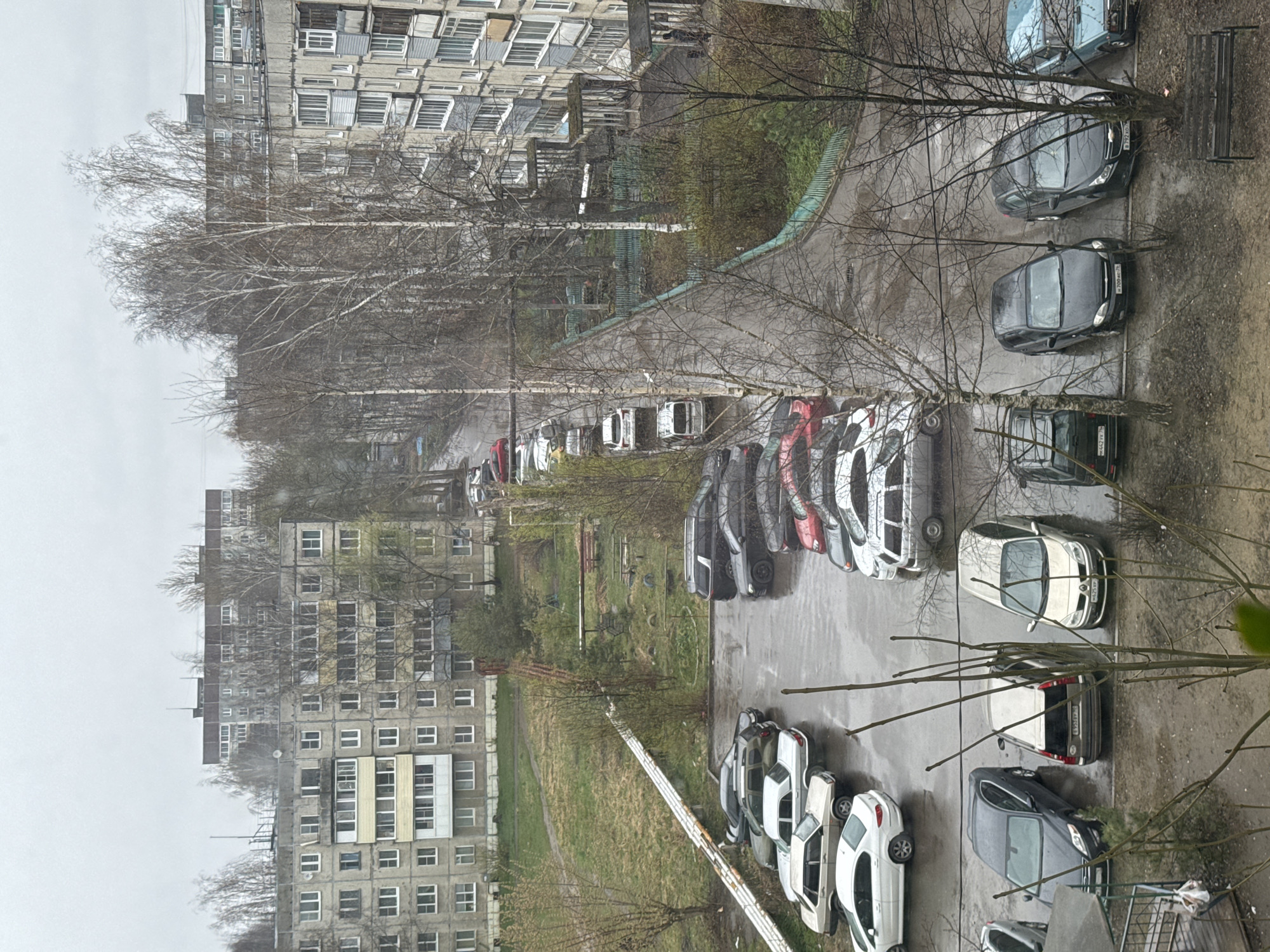 The image shows an aerial view of a residential area in Ново-Никольское, Russia. Several gray apartment buildings dominate the landscape. A parking lot is visible, containing a variety of cars, mostly in shades of white, gray, and black. Bare trees are interspersed between the buildings and the parking lot. The ground appears wet, suggesting recent rain. A patch of green grass is also visible. The sky is overcast and gray.