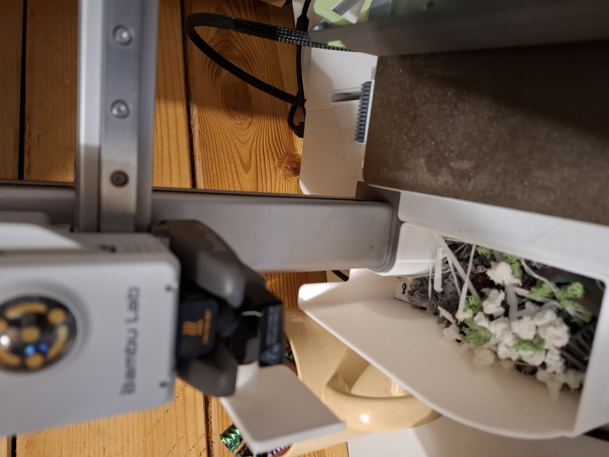 The image displays a close-up, slightly angled view of a white 3D printer, identified by the "Bambu Lab" logo visible on its upper casing. The printer's print head, a dark grey component with a yellow symbol, is positioned above a white collection bin. This bin contains various small, colorful objects, appearing to be remnants or debris from the printing process, including white and green floral-like shapes and other unidentifiable small items. To the left of the collection bin, a beige pitcher is partially visible. The background consists of a wooden plank wall, suggesting an indoor setting. A black cable, likely for power or data, snakes across the wooden surface behind the printer. The overall scene is utilitarian, focusing on the components of the 3D printer and its immediate surroundings. The location in the city of Tyumen, Russia, is not discernible from the visual information presented in the image.