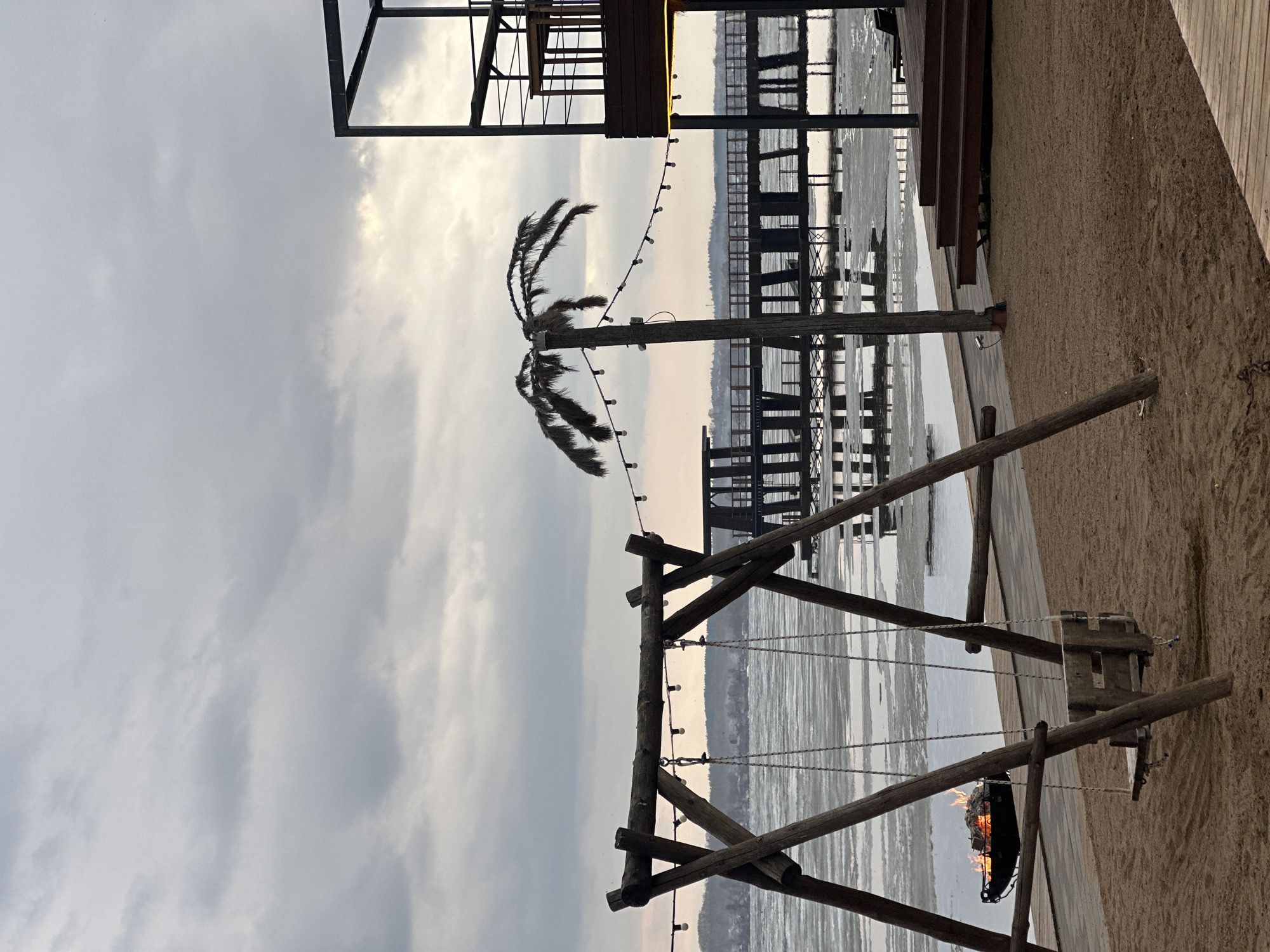 The image presents a scene at a waterside location in Votkinsk, Russia. A decorative wooden swing set stands prominently in the foreground, with a fire burning in its seat. A palm tree, adorned with string lights, rises in the background near a boardwalk or pier extending over the water. Additional building structures are visible in the top of the image. The sky is overcast, suggesting either dawn or dusk. The scene is characterized by a contrast between natural elements and constructed infrastructure, creating an atmospheric setting.