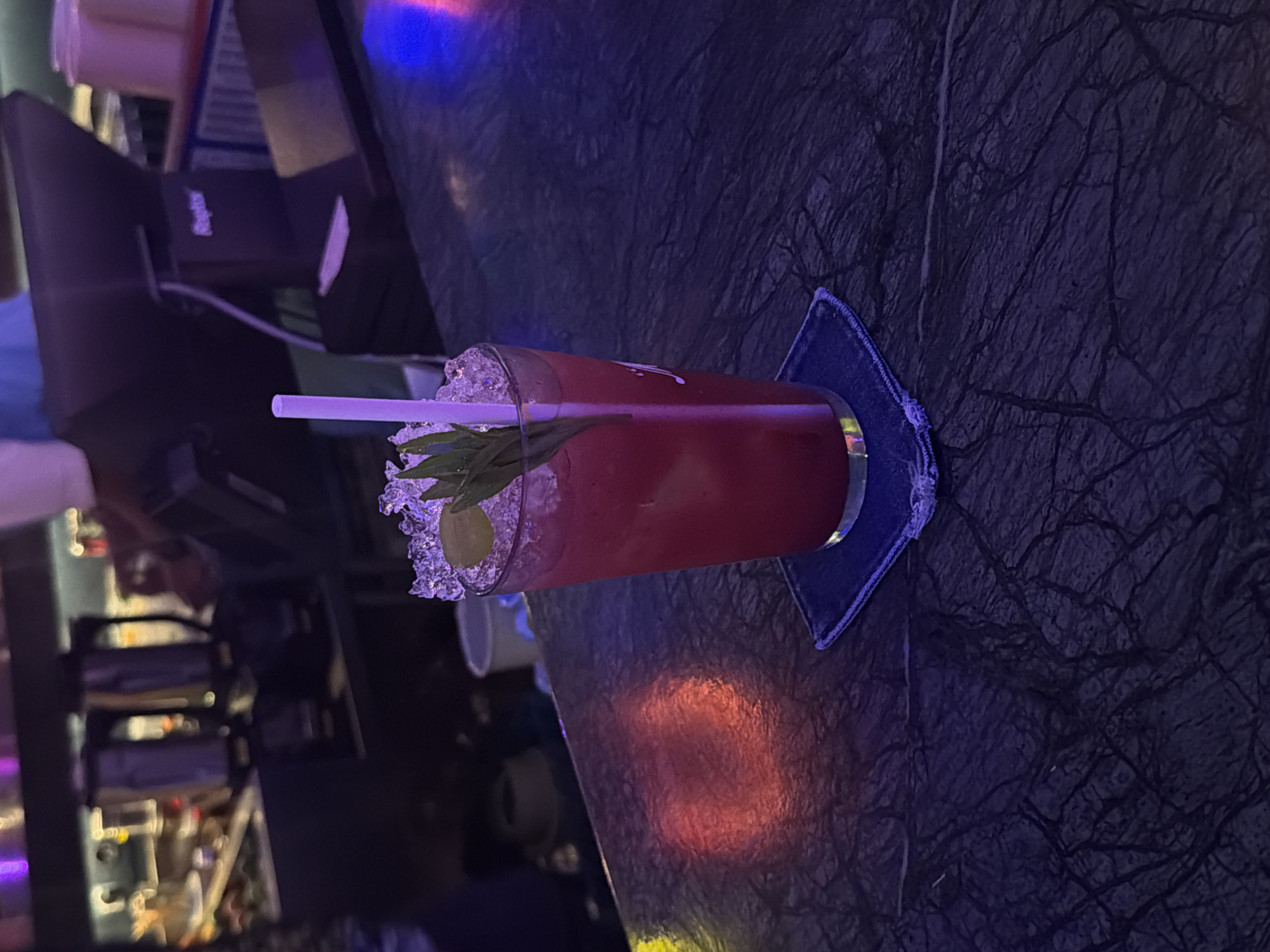 A close-up, eye-level shot shows a vibrant pink cocktail with ice, a sprig of mint, and a light purple straw in a glass. The glass rests on a blue coaster on a dark, textured countertop. The background is blurred, revealing a bar area with shelves holding various bottles and equipment. A point-of-sale system is visible on a counter. Red and blue lights cast reflections on the countertop. The image appears to be taken indoors at night, possibly in a bar or restaurant setting. The location is identified as North Kuta, Indonesia.