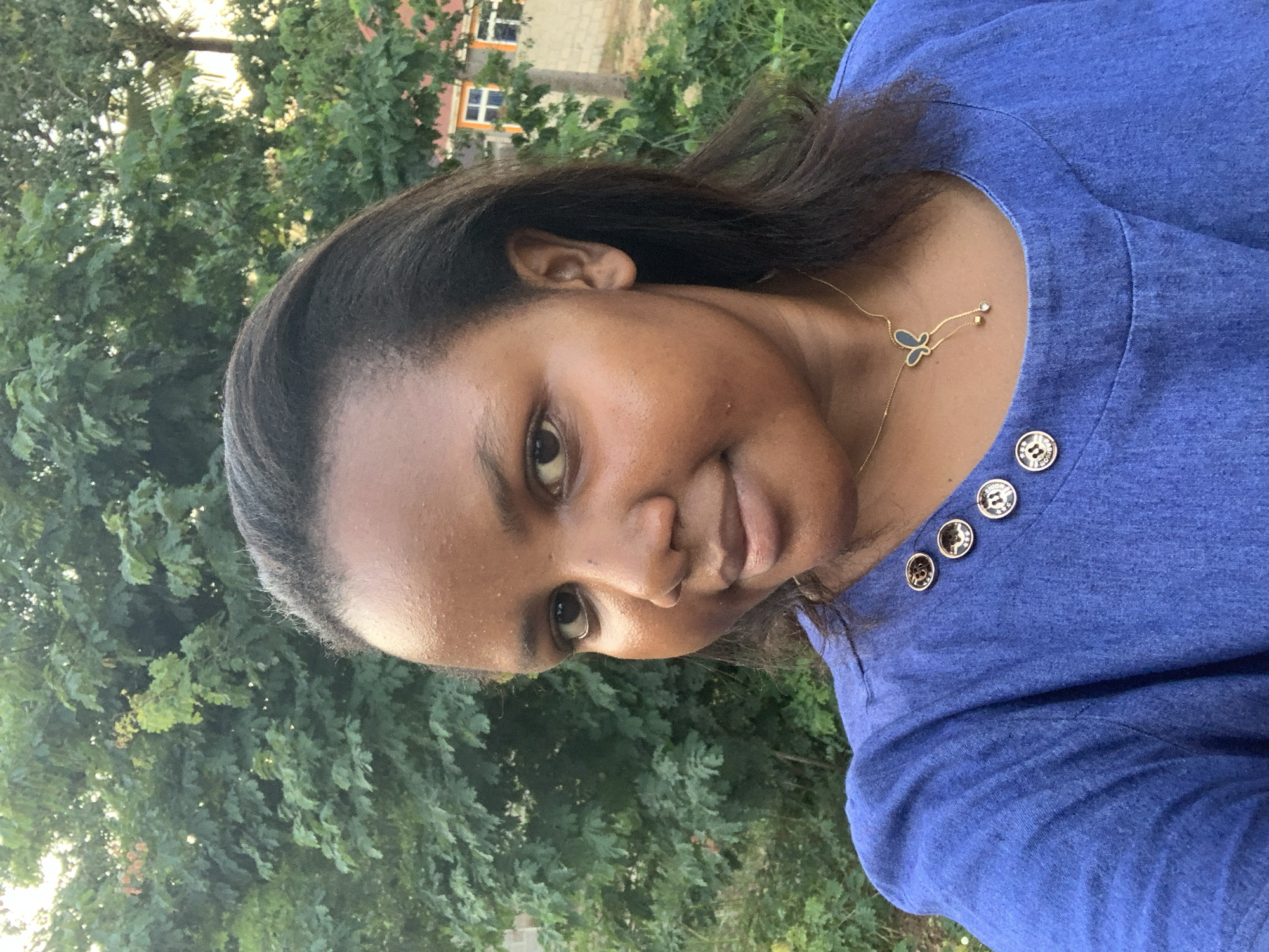 A medium, outdoor shot depicts a young Black woman wearing a blue button-down shirt and a butterfly necklace. The background consists of green trees and a building. The location is implied to be Dar es-Salaam, Tanzania.
