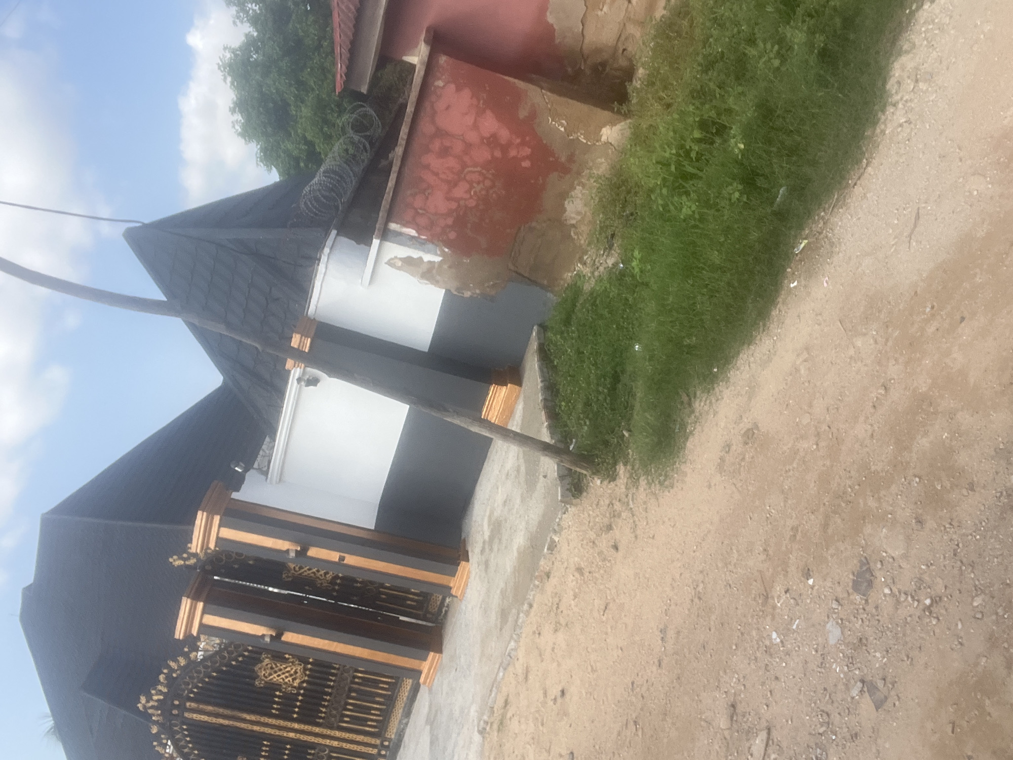 The image depicts a street view in Jalingo, Nigeria, focusing on the exterior of a residential property. The property features a prominent black-tiled roof with a steep pitch, supported by dark grey and white walls. A decorative black and gold metal gate stands at the entrance, flanked by pillars adorned with dark grey and gold accents. Adjacent to the main house, a separate structure displays peeling red paint and visible signs of disrepair, with barbed wire coiled along its top. The ground in front of the property is a dirt path mixed with gravel, with a strip of green grass and foliage bordering the road. A thin, wooden pole stands vertically in the foreground, partially obstructing the view of the building. The sky above is partly cloudy. No people or animals are visible in the scene.