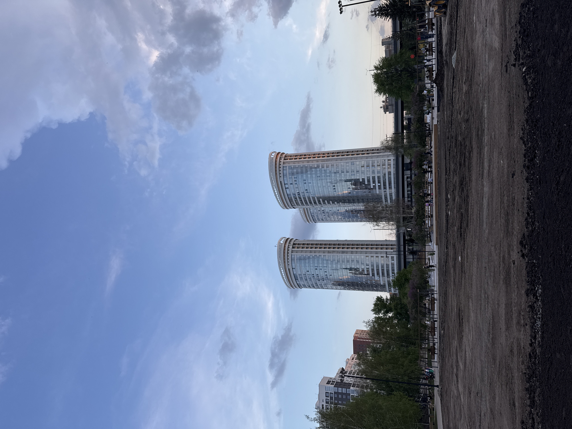 The image shows two modern cylindrical buildings with glass facades in Novosibirsk, Russia. The buildings appear to be high-rise residential or commercial structures. A large open area is in the foreground, appearing to be a construction site or a newly developed public space, with dirt and gravel covering the ground. In the background, there are other buildings and some trees and street lamps. The sky is visible, with clouds scattered across a blue expanse. The scene is illuminated by daylight, suggesting it is daytime. There is evidence of paving work and some construction equipment is apparent. The presence of trees, benches, and street lamps hint at the potential to be a park or urban plaza. There is minimal human activity, just a few individuals standing or sitting on benches in the park.