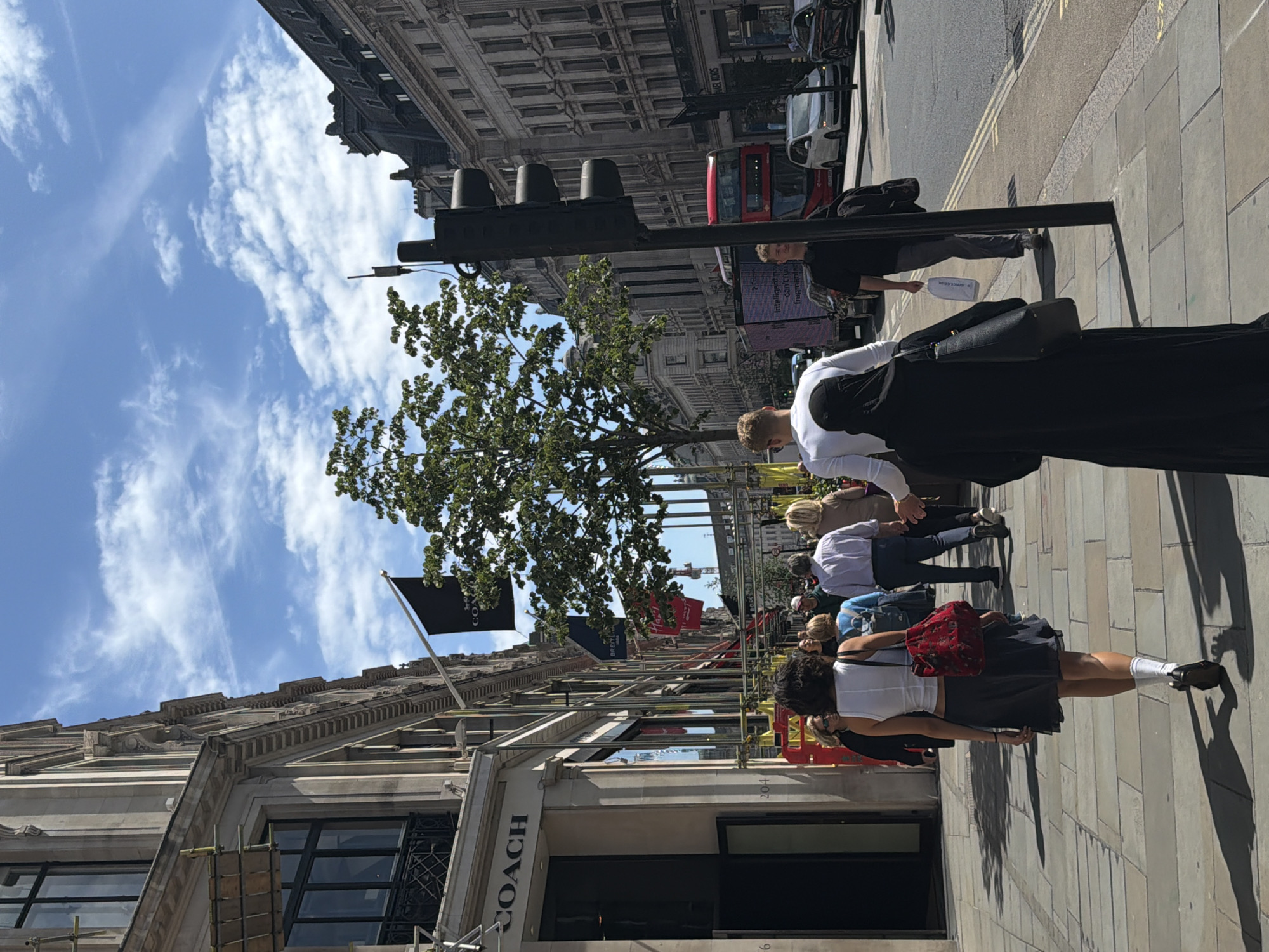 A photograph depicts a street scene on a sunny day in London, United Kingdom. The street features tall buildings with ornate facades, and scaffolding is visible along the lower portion of one building. A "COACH" store is visible. Pedestrians, including individuals of various ages and clothing styles, are walking along the sidewalk. A black traffic light fixture with three signals is positioned along the street. A red double-decker bus and other vehicles are partially visible in the background. Banners and flags hang from the buildings. The sky is blue with scattered clouds.