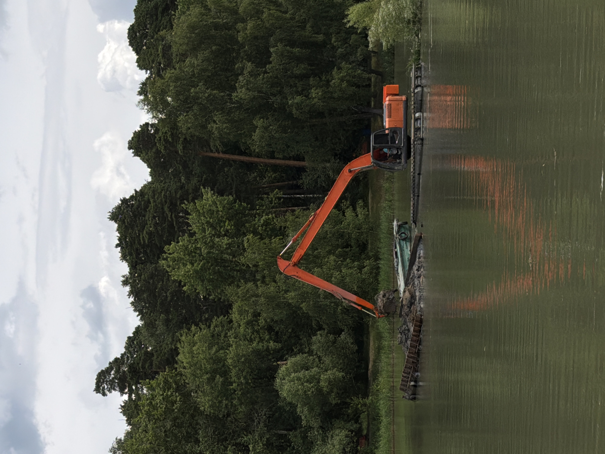 An orange excavator is operating from a floating platform on a body of water near a wooded area. The excavator's arm is raised and appears to be scooping or depositing material. The platform itself is also carrying a quantity of dirt and possibly debris, with a small green boat visible alongside it. The background is dominated by a dense forest, with a cloudy sky overhead. The scene suggests ongoing construction or dredging work. The location is the city of Bykovo, Russia.