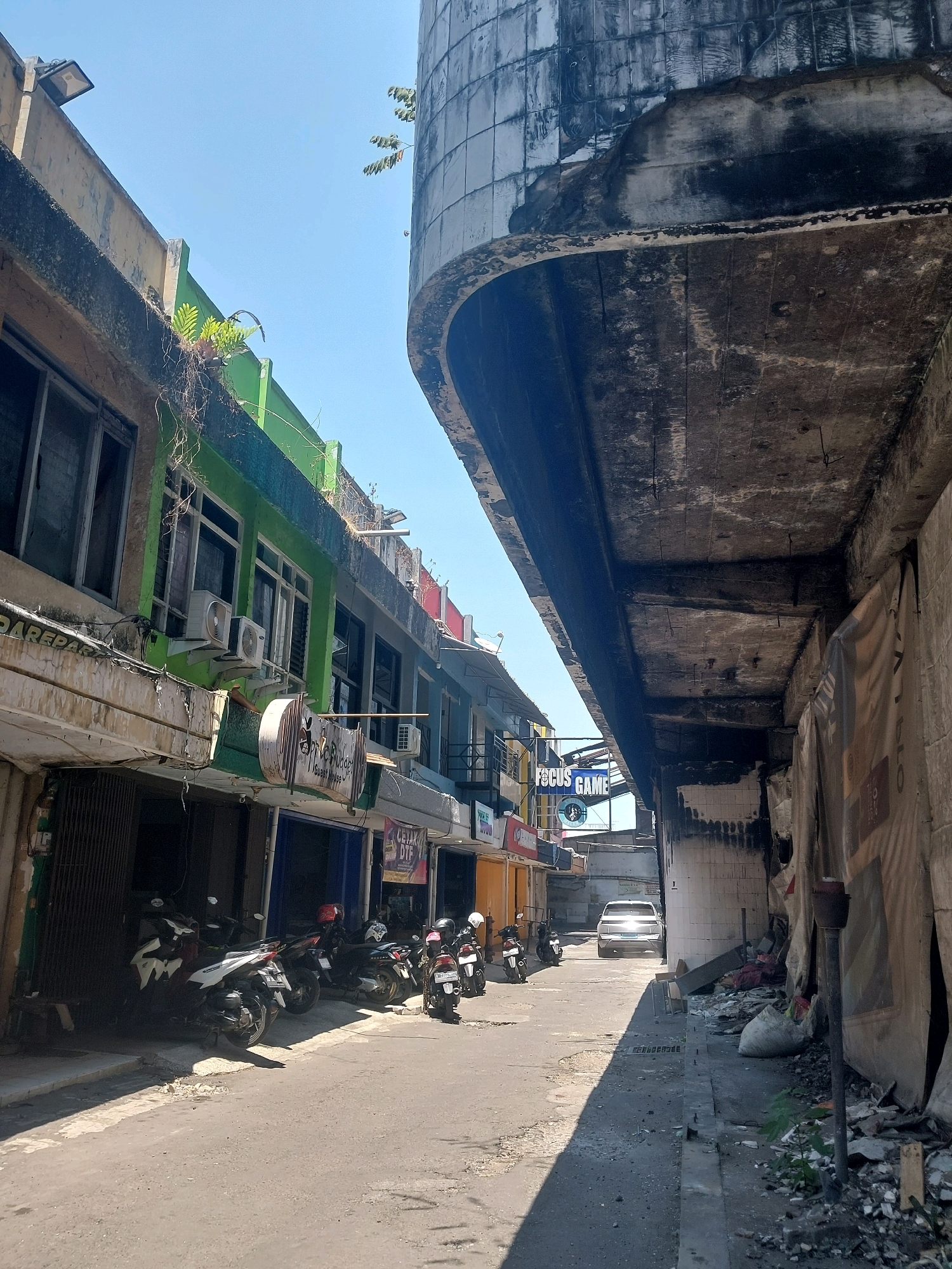 The image captures a narrow street scene in Malang, Indonesia, lined with commercial buildings and parked motorcycles. On the left, a row of shops features storefronts with metal grates and visible air conditioning units. Some of the buildings are painted in vibrant colors, like green and yellow, with plants growing along the upper ledges. Signs are visible, including one that reads "FOCUS GAME". The street itself is paved, with the sun casting sharp shadows from the buildings. On the right side of the street, a large, curved concrete structure dominates, showing signs of wear and decay, with visible exposed rebar and water damage. Debris is piled up along the base of this structure. A white car is visible further down the street, moving away from the camera. The sky is clear and blue, suggesting a sunny day.