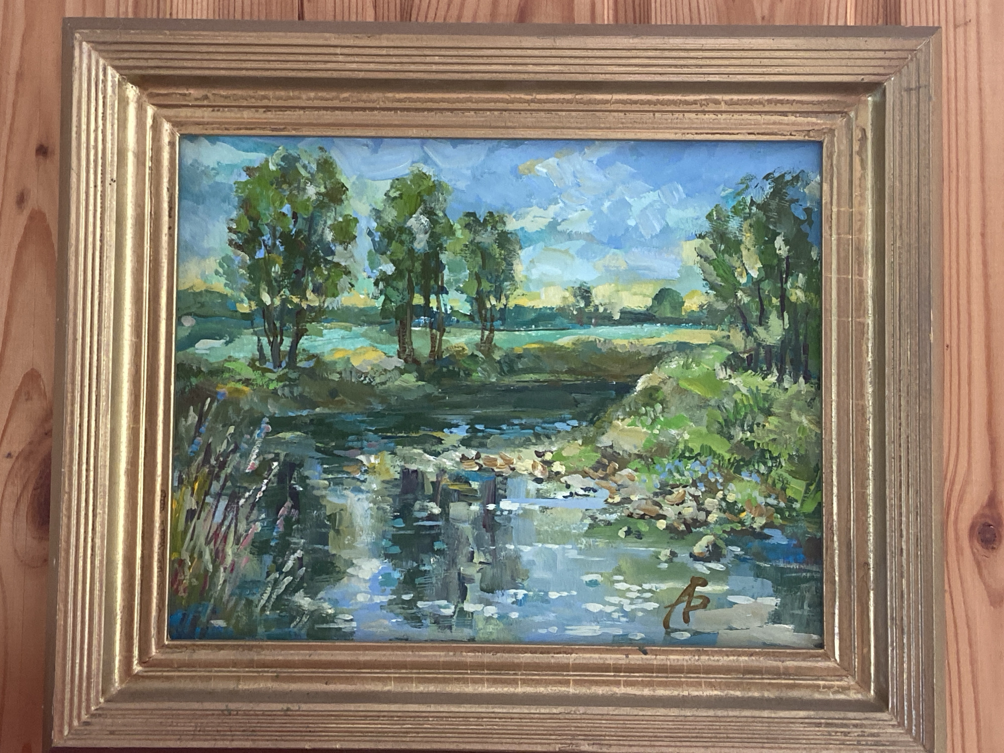 This is an oil painting of a tranquil river scene, framed in a golden, ridged frame. The painting depicts a body of water with reflections of trees and sky. Along the riverbanks, there is lush greenery and scattered rocks. Several trees with full canopies are visible, contributing to the natural landscape. The sky is rendered with a mix of blues and white clouds, suggesting a bright, possibly partly cloudy day. In the lower right corner of the painting, a stylized signature in a darker color is present. The painting is hung on a wooden wall. There is no identifiable connection to the city of Naberezhnye Chelny, Russia.