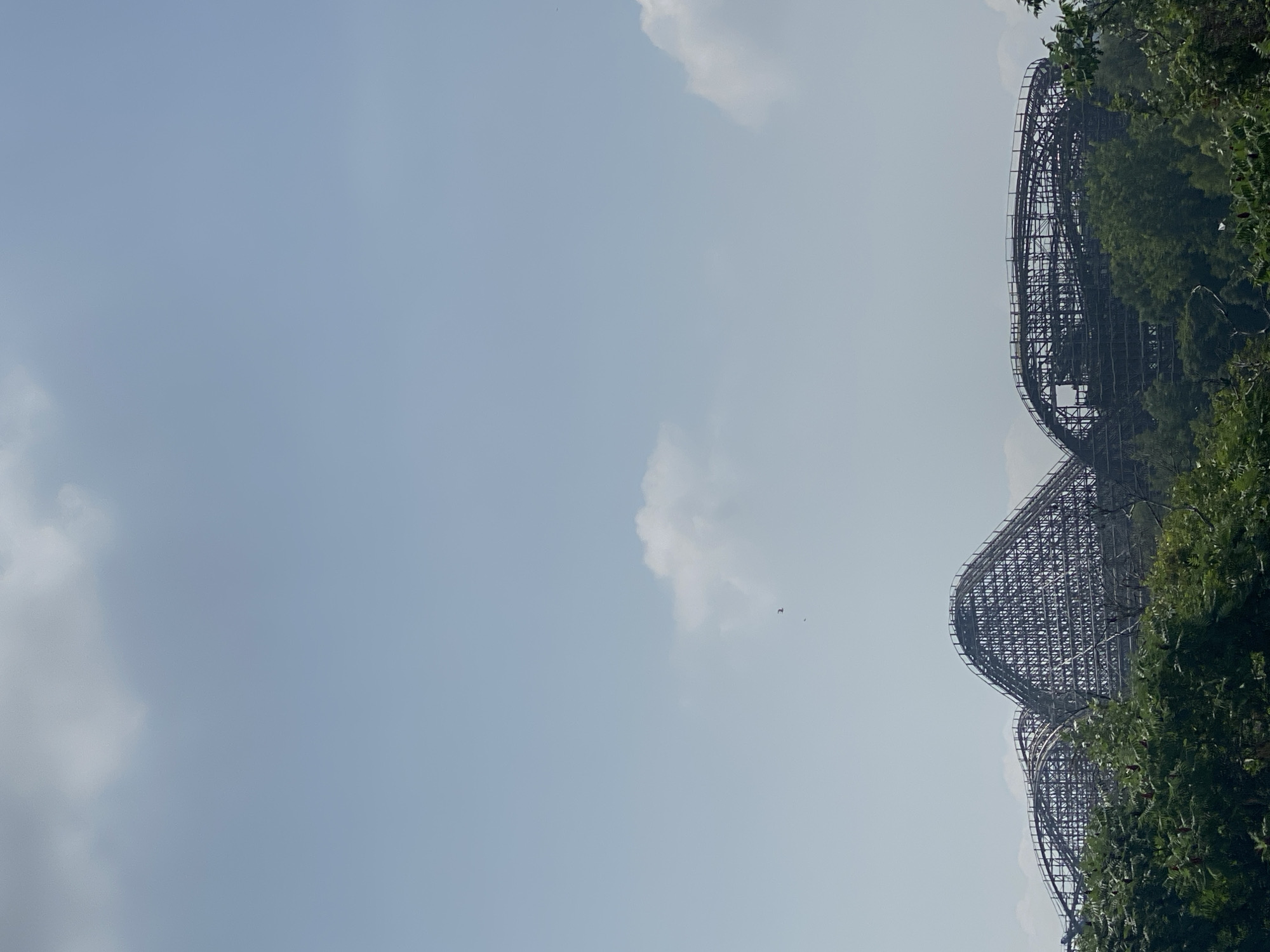 The image shows a large wooden roller coaster with a complex track structure, partially obscured by green trees in the foreground. The roller coaster has multiple hills and drops, with a prominent peak on the left side of the frame. The sky is a pale blue with scattered white clouds, and there are two small dark specks, possibly birds, in the sky above the roller coaster. The scene appears to be an outdoor amusement park. The image does not provide any specific clues to its location in Montreal, Canada.