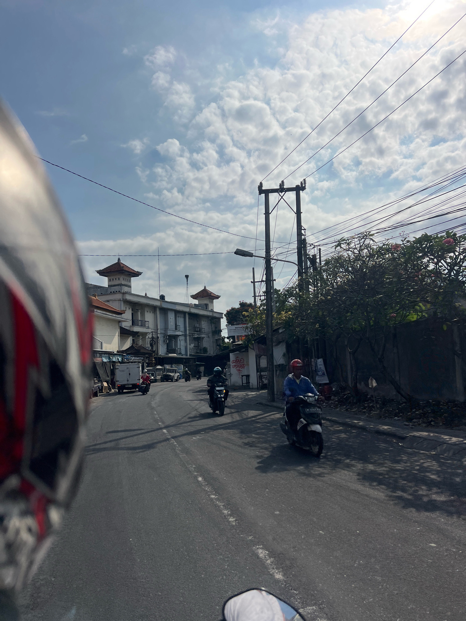 This is a POV shot from a motorcycle, looking down a street in Kuta, Indonesia. The street is paved and appears to be moderately busy with traffic. In the foreground, the rider's helmet is partially visible on the left side of the frame. A person on a scooter wearing a blue shirt and a red helmet is riding in the right lane, with a few other scooters and a white van further down the road. Buildings line the street, with one prominent building in the mid-ground featuring a distinctive Balinese-style roof. Power lines crisscross the sky, which is partly cloudy with the sun shining through. Trees with pink flowers are visible on the right side of the street. The overall scene depicts a typical day on a street in a bustling Indonesian city.