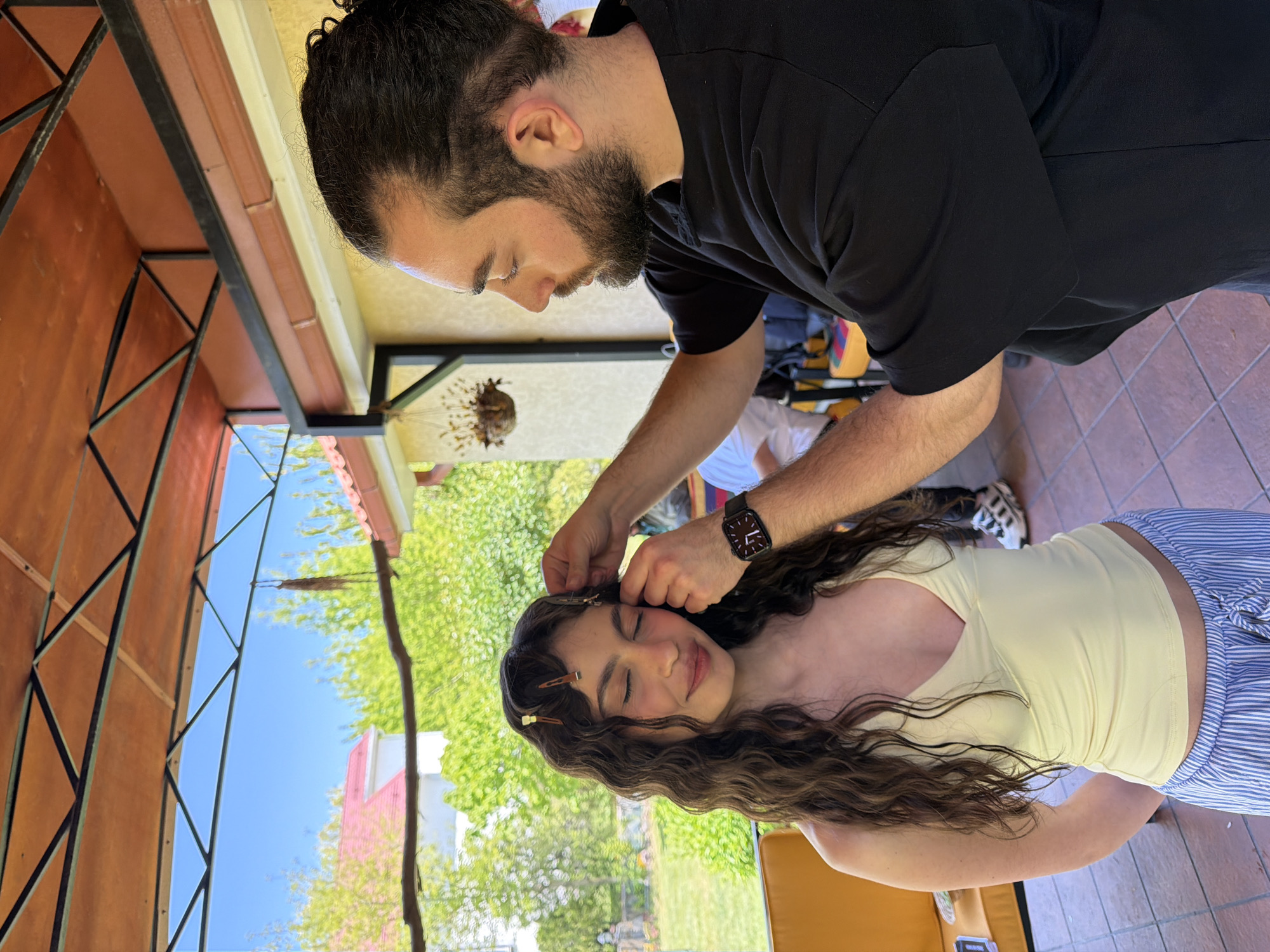 A medium shot shows a man tending to the hair of a young woman outdoors. The man has dark hair pulled back in a bun and a beard; he wears a black t-shirt and a watch with a dark band. The woman has curly brown hair, a light yellow tank top, and light blue and white striped pants. She sits with her eyes closed, and the man's hands work in her hair, securing it with bobby pins. The scene takes place under a metal frame structure, possibly a patio or outdoor salon. Behind the metal frame, there are green trees, a blue sky, and a glimpse of a pink building. The background includes glimpses of tiled flooring and possibly equipment or a work area. The location is likely Yerevan, Armenia, but there's no definitive information in the photo to confirm that.