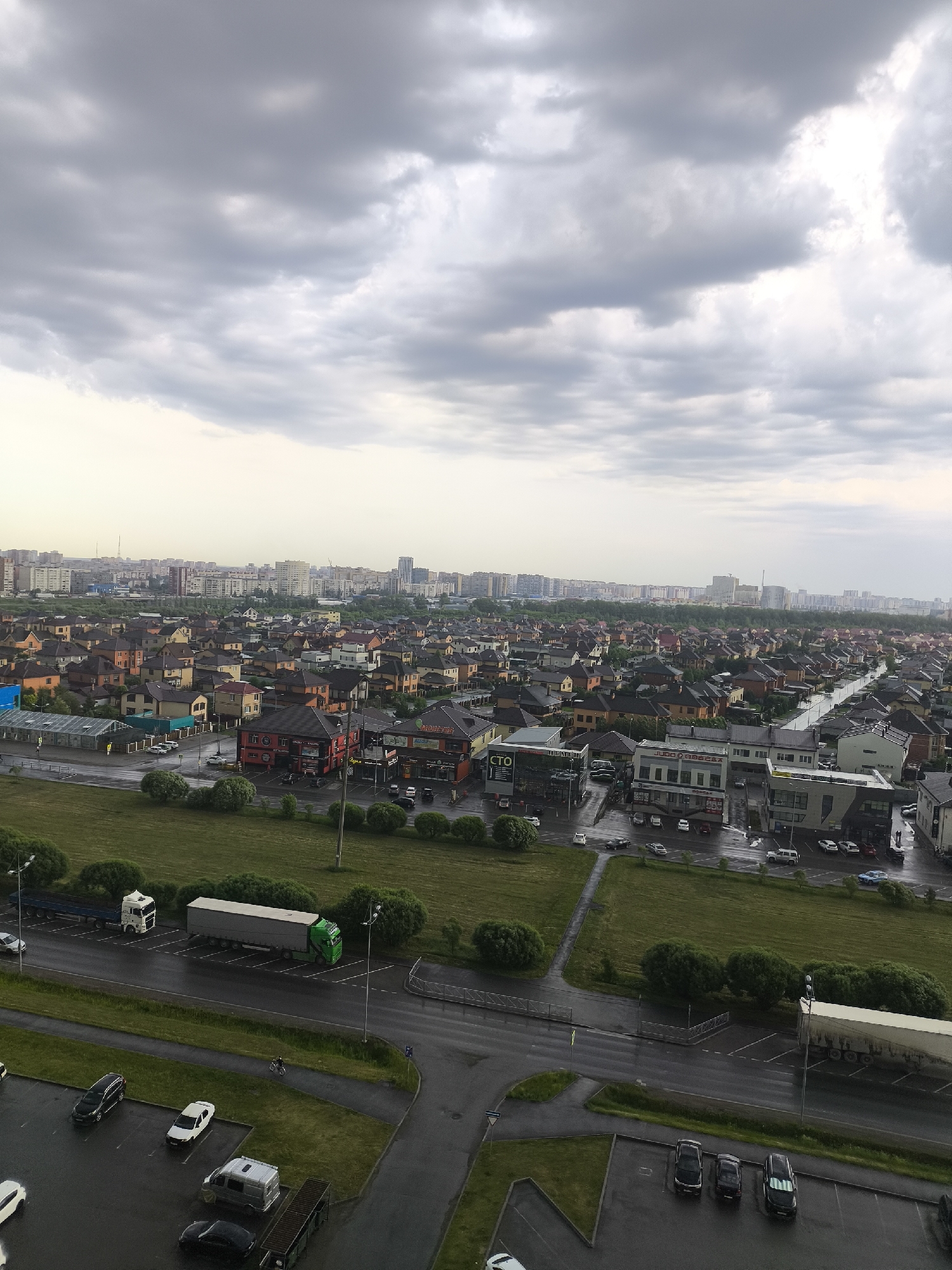 The image is an elevated view of a residential and commercial area in Tyumen, Russia, taken on an overcast day.
**Environment:** The sky is filled with gray clouds, suggesting rain or recent rainfall. The ground is wet, indicating a recent rain event. There are green grassy areas and trimmed bushes separating the road from the commercial buildings and residences.
**Location:** The image shows a mixed urban landscape of low-rise buildings, detached houses, and a cityscape with taller buildings in the distance. Commercial buildings line the street, including a building with signage "CTO." There are residential homes with dark roofs behind the commercial zone.
**Objects:**
* Roads and parking areas with parked vehicles.
* A large truck with a green cab and a white trailer is parked on the road.
* Various cars are parked in parking lots, including a white van and several dark-colored sedans.
* Buildings of varying architectural styles: residential houses with pitched roofs, commercial buildings with storefronts.
**People:** There is a cyclist on the sidewalk.
**Actions/Interactions:** The scene appears calm and still, with no significant activity taking place. The presence of parked vehicles suggests a functional, occupied area.
**Overall Impression:** The image portrays a typical urban landscape in Russia, specifically Tyumen, with a mix of residential and commercial zones under an overcast sky.