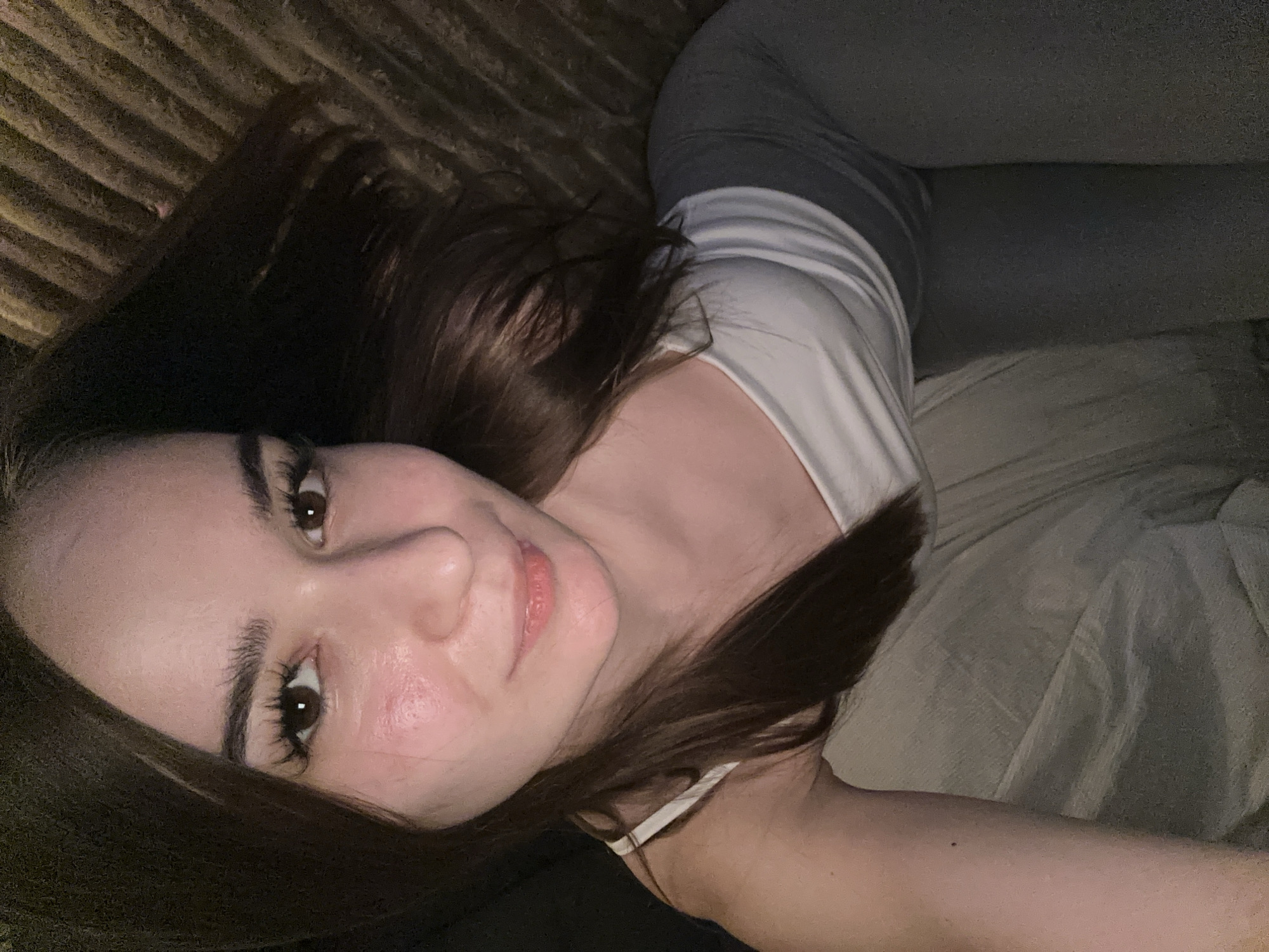 A woman is lying down and taking a selfie. She has long, dark hair, dark eyes, and full lips. She is wearing a white tank top and gray leggings. The background is blurry, but it appears to be a bed or couch. The lighting is dim, and there is a reddish tint to the image.