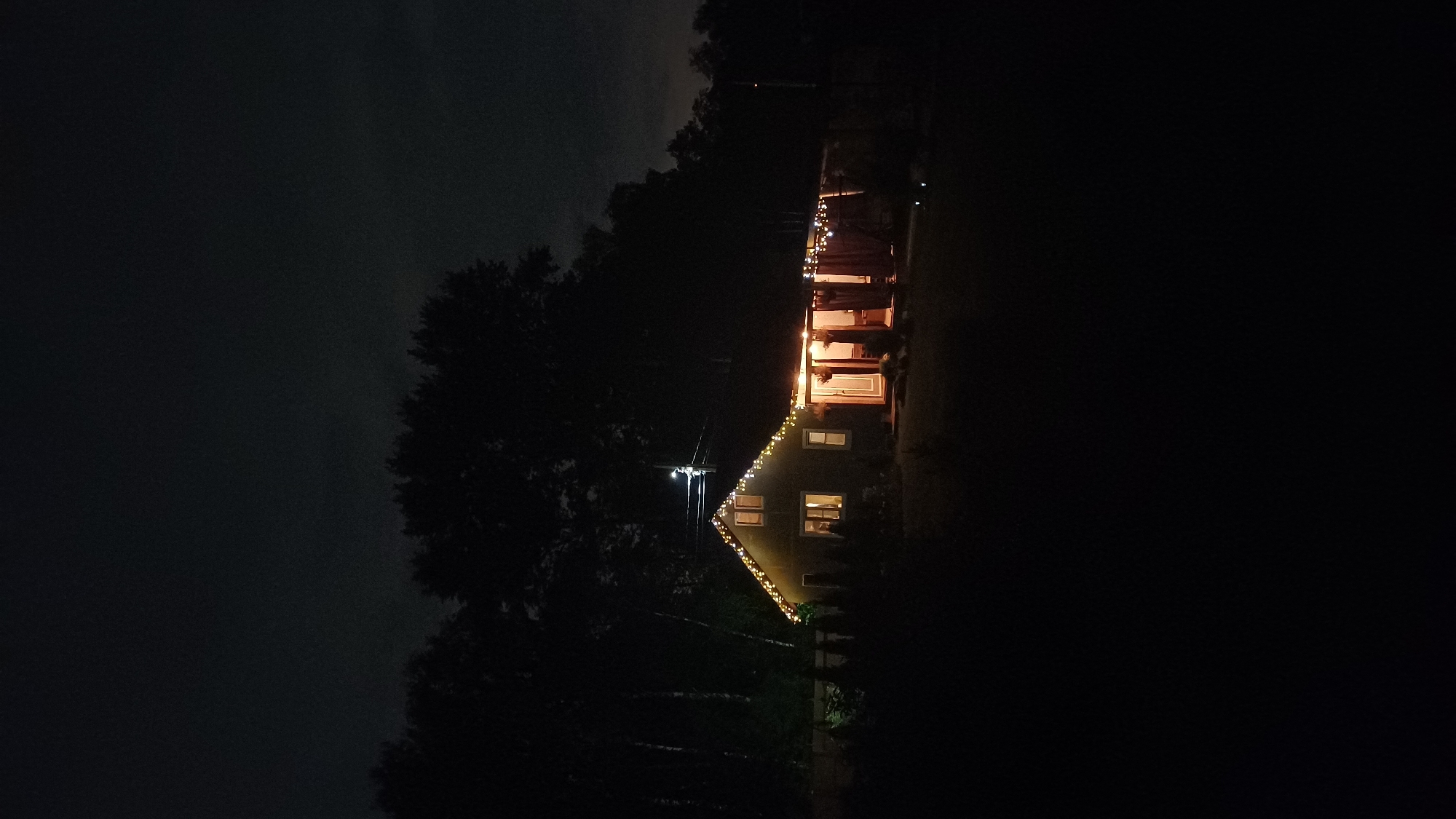 At night in Timokhovo, Russia, a house is illuminated with lights strung along its roofline and porch. Trees are visible in the foreground and surrounding the house. The sky is dark and cloudy.