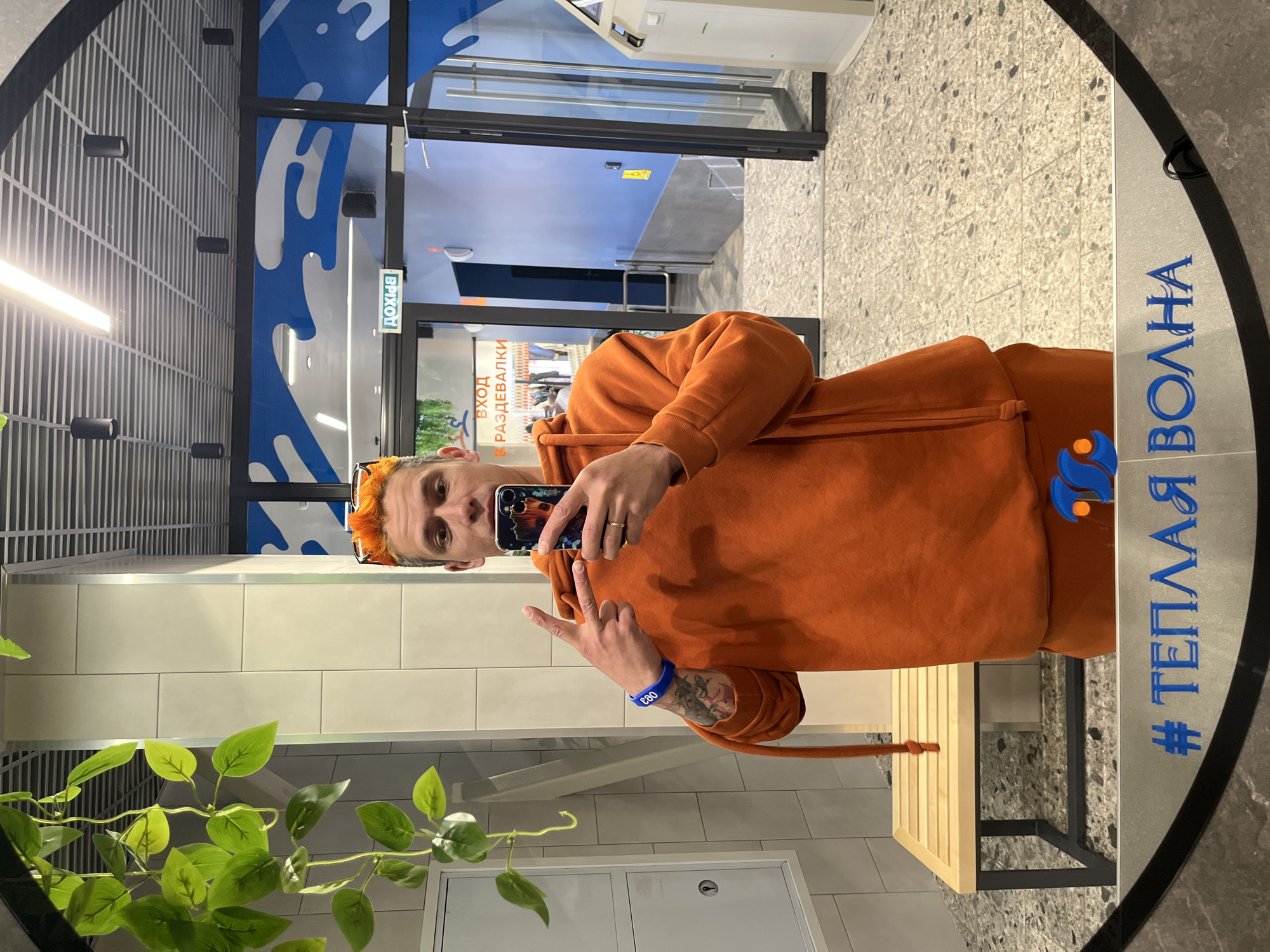 A man with orange hair is taking a selfie in a mirror. He is wearing an orange hoodie and has a blue wristband on his left wrist. He is holding a phone in his right hand and making a peace sign with his fingers.
The mirror has the words "# ТЕПЛАЯ ВОЛНА" written on it in blue.
The location appears to be an indoor facility, potentially a water park or recreational center. Behind the man, there is a doorway with a sign in Russian, indicating the entrance to the dressing rooms. There are also decorative blue wave patterns on the walls.
Based on the mirror and general aesthetic, this image was likely taken in the "Teplaya Volna" water park, located in Votkinsk, Russia.